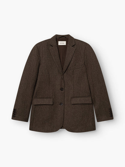 Der Blazer „Karoline Braun“ ist ein taillierter Blazer mit fallendem Revers, zwei Knöpfen, einer Brusttasche und zwei Pattentaschen vorne. Er ist aus weichem Baumwollflanell gefertigt, was ihm eine edle Optik verleiht. Das Modell wird in Portugal hergestellt und hier flach auf einem weißen Hintergrund präsentiert.