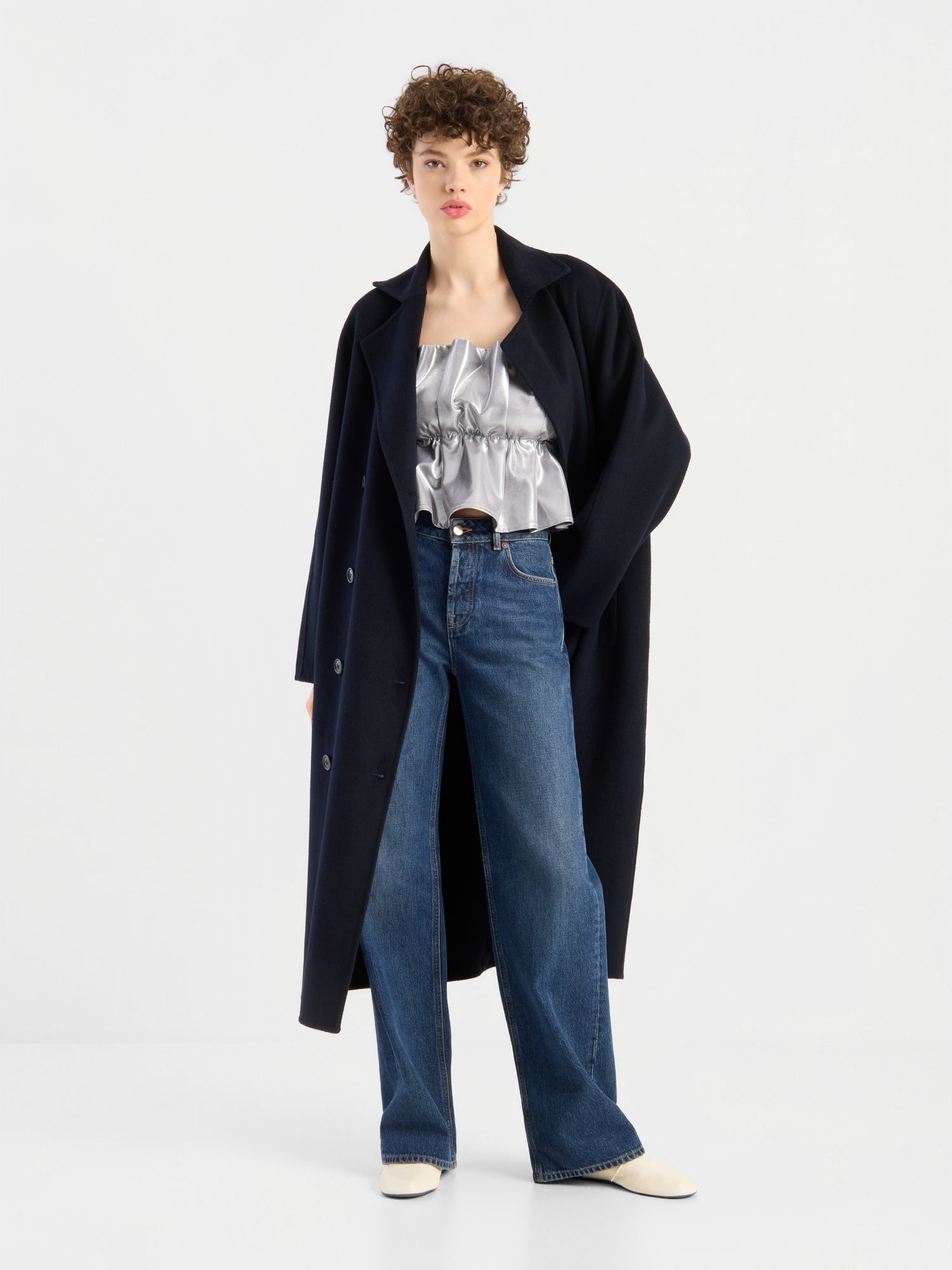 Eine Person, die einen langen schwarzen Mantel über einem glänzenden silbernen „Pleat Top Silber“ mit Rüschenverzierungen, einer blauen Jeans mit weitem Bein und weißen Schuhen trägt, steht vor einem schlichten weißen Hintergrund.