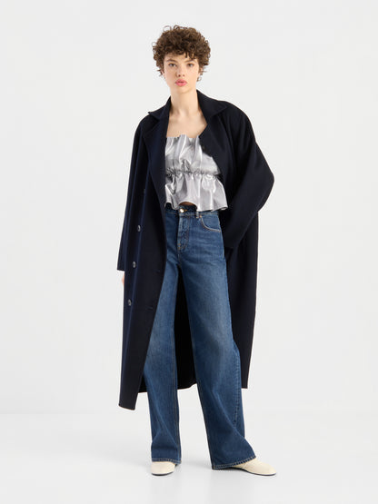 Eine Person, die einen langen schwarzen Mantel über einem glänzenden silbernen „Pleat Top Silber“ mit Rüschenverzierungen, einer blauen Jeans mit weitem Bein und weißen Schuhen trägt, steht vor einem schlichten weißen Hintergrund.