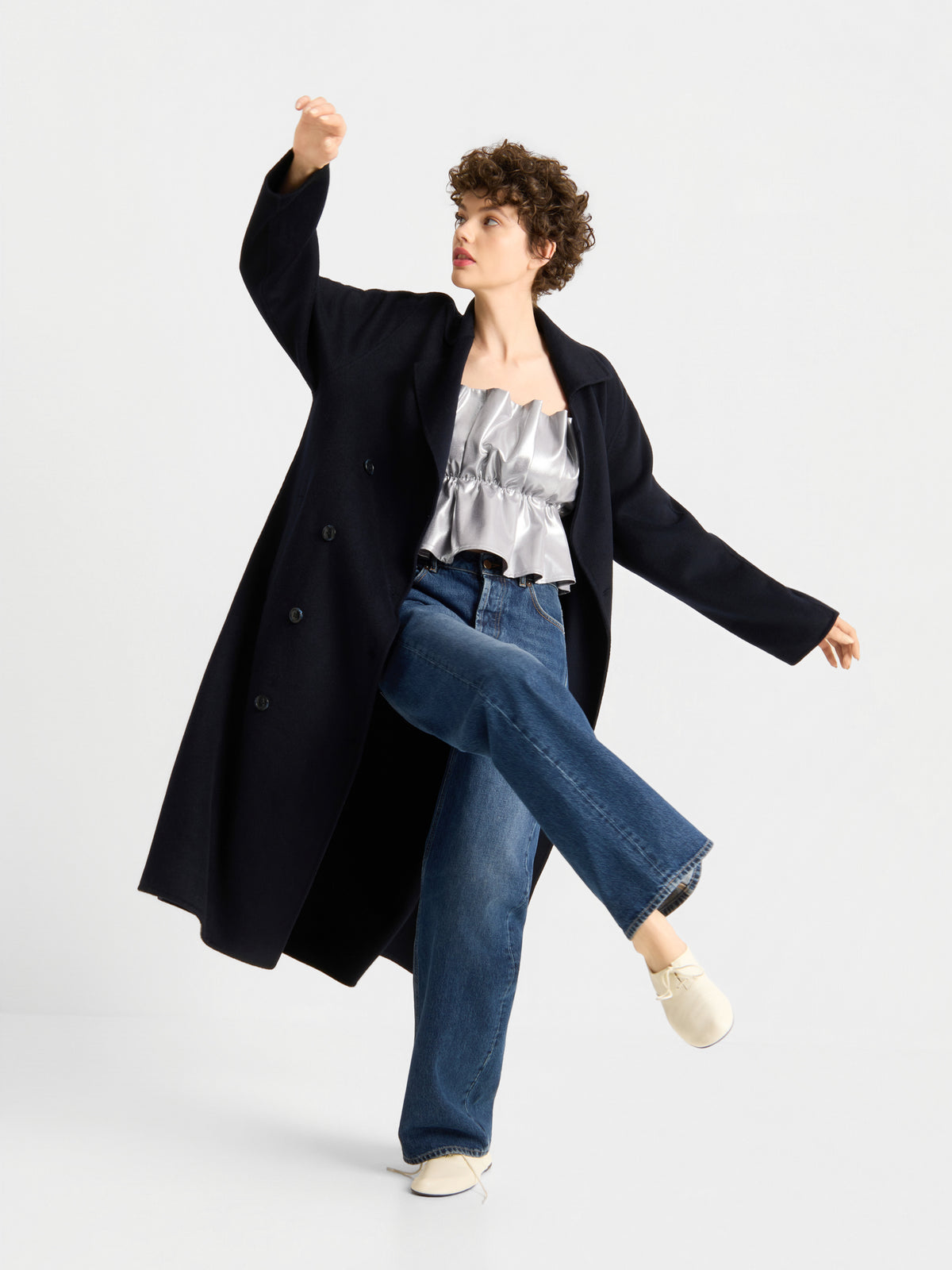 Eine Person, die einen langen schwarzen Mantel, das „Pleat Top Silber“, eine blaue Jeans und weiße Schuhe trägt, posiert dynamisch vor einem schlichten weißen Hintergrund.