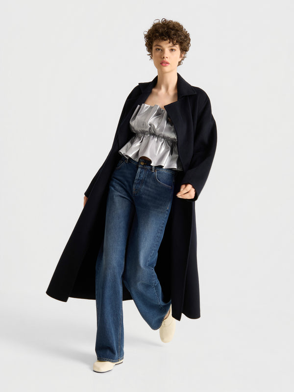 Eine Person, die einen langen schwarzen Mantel, das Oberteil „Shine in Silver“ in Metallic-Optik, blaue Jeans und weiße Schuhe trägt, steht vor einem schlichten, hellen Hintergrund.