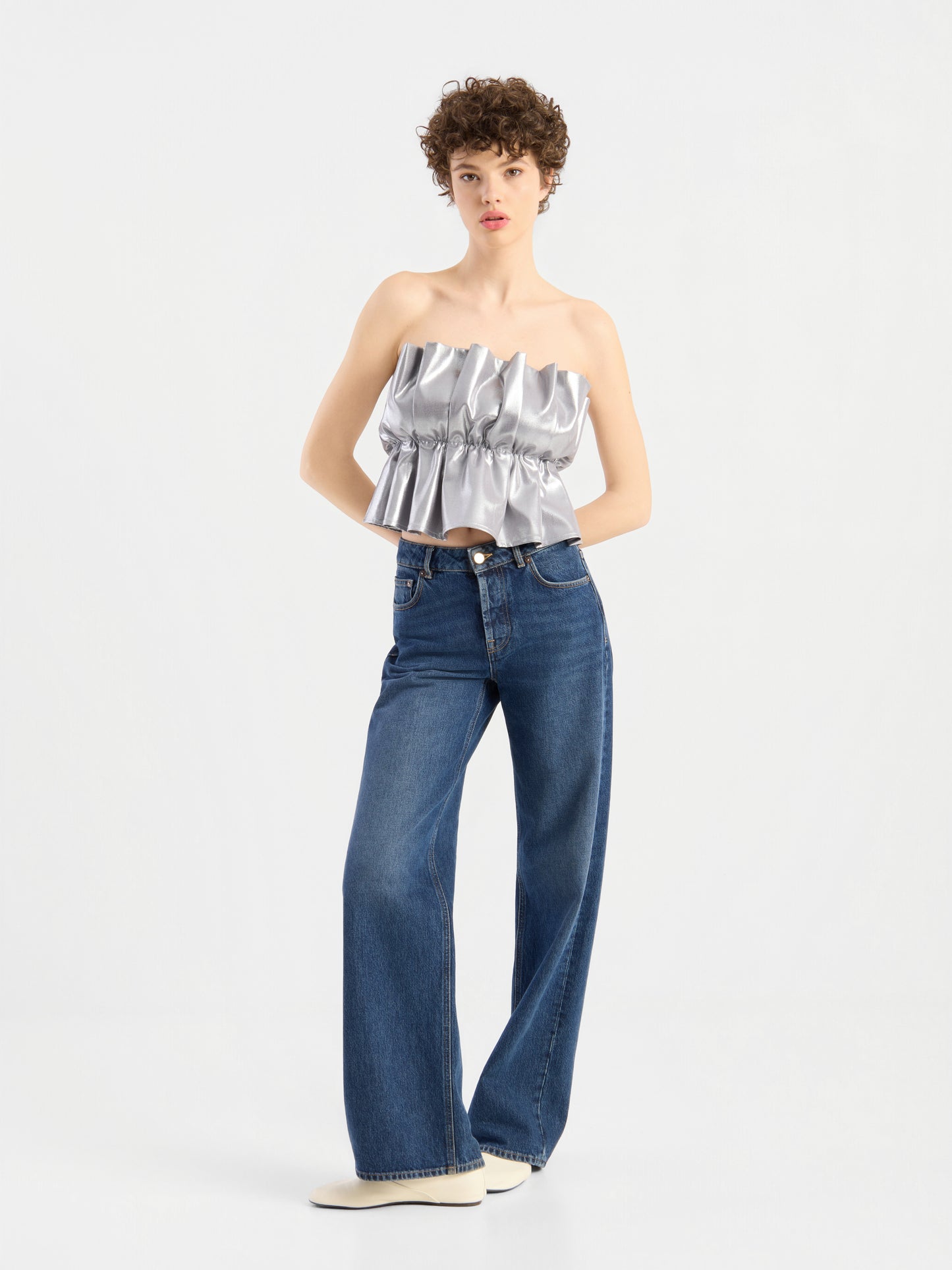Eine Person mit kurzen Locken trägt ein „Pleat Top Silber“ mit Rüschen, eine blaue Jeans mit weitem Bein und weiße Schuhe und steht vor einem schlichten weißen Hintergrund.