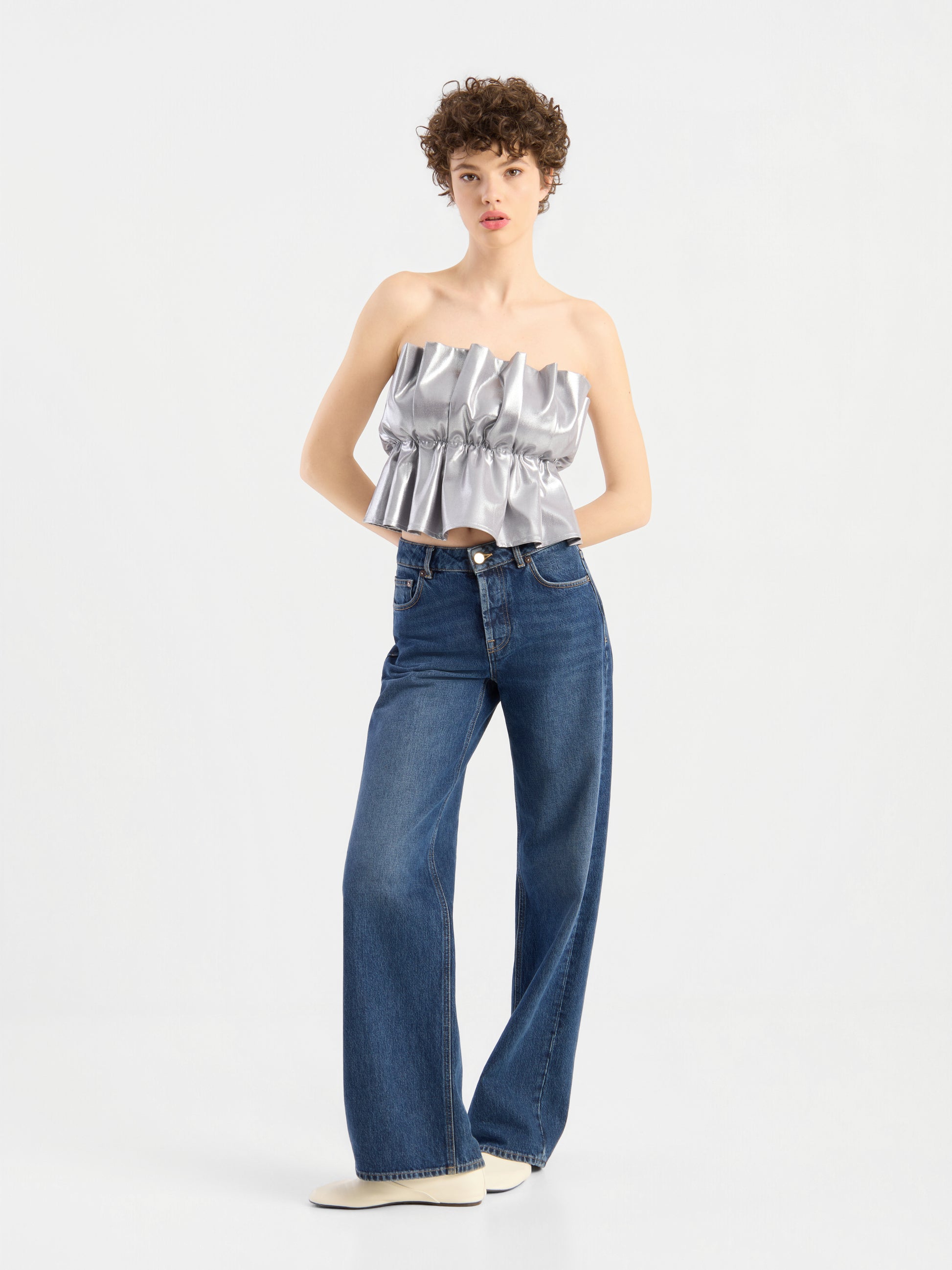 Eine Person mit kurzen Locken trägt ein „Pleat Top Silber“ mit Rüschen, eine blaue Jeans mit weitem Bein und weiße Schuhe und steht vor einem schlichten weißen Hintergrund.