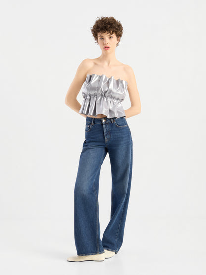 Eine Person mit kurzen Locken trägt ein „Pleat Top Silber“ mit Rüschen, eine blaue Jeans mit weitem Bein und weiße Schuhe und steht vor einem schlichten weißen Hintergrund.