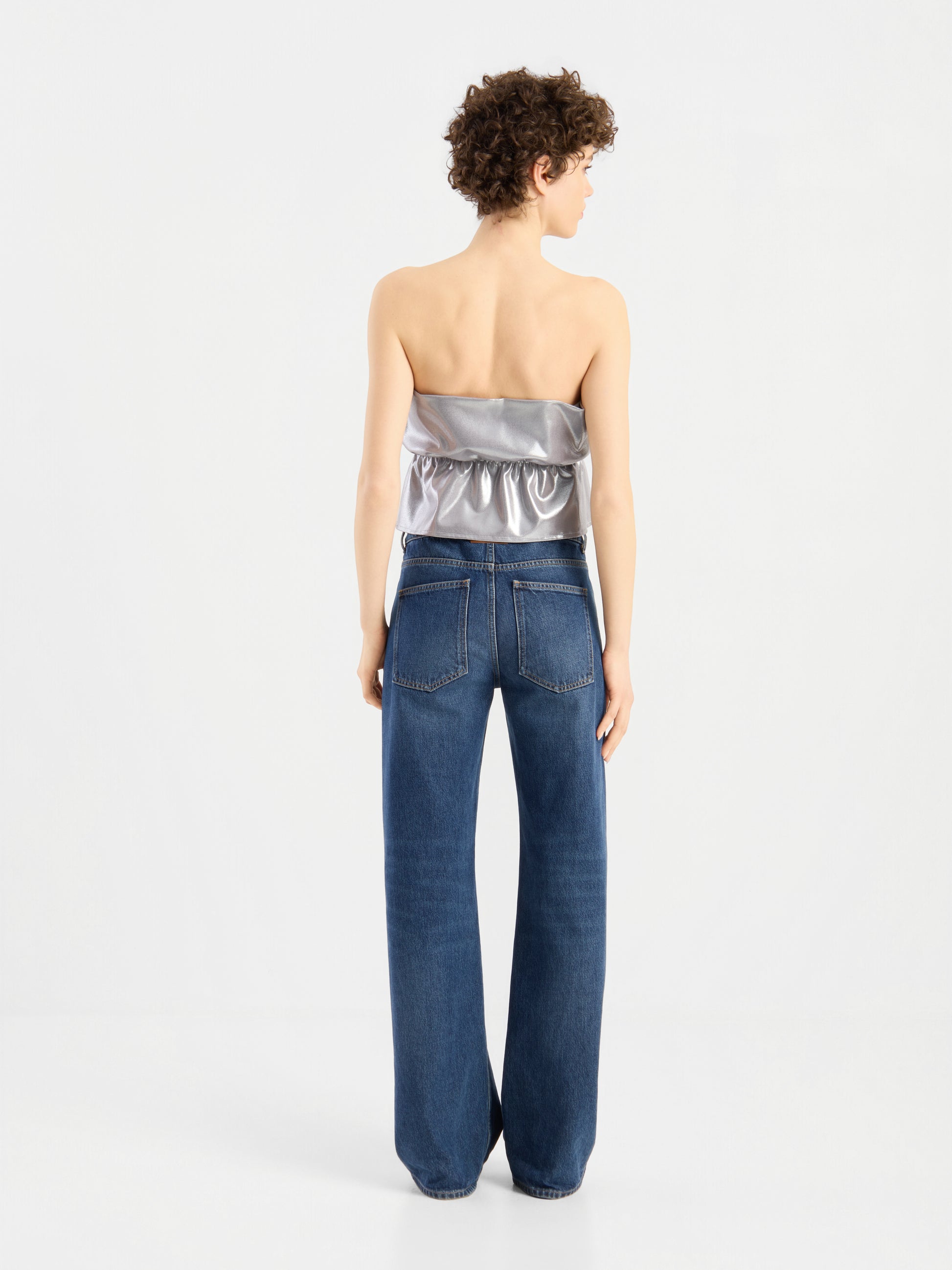 Eine Person mit kurzen Locken steht mit dem Rücken zur Kamera und trägt das „Pleat Top Silber“ sowie eine lockere blaue Jeans vor einem schlichten weißen Hintergrund.