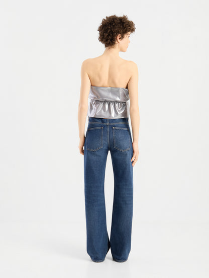 Eine Person mit kurzen Locken steht mit dem Rücken zur Kamera und trägt das „Pleat Top Silber“ sowie eine lockere blaue Jeans vor einem schlichten weißen Hintergrund.