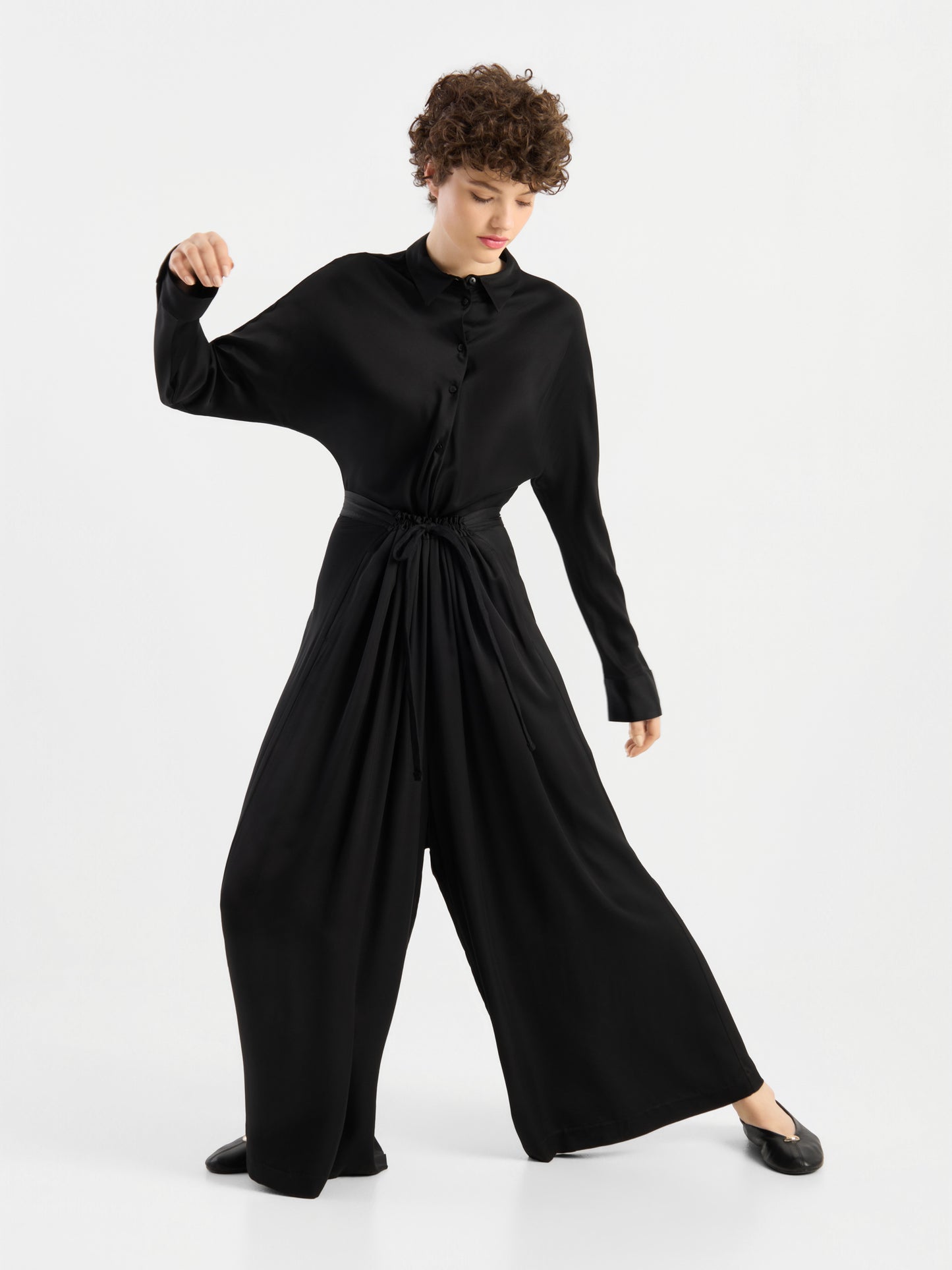 Eine Person mit kurzen Locken, die die Bluse „Kaia Schwarz“ – eine locker sitzende schwarze Bluse – trägt, kombiniert mit schwarzen Schuhen, steht vor einem weißen Hintergrund in einer dynamischen Pose, wobei ein Arm angewinkelt und der andere ausgestreckt ist. Die Bluse wird in Portugal hergestellt und zeugt von hochwertiger Handwerkskunst und zeitgemäßem Stil.