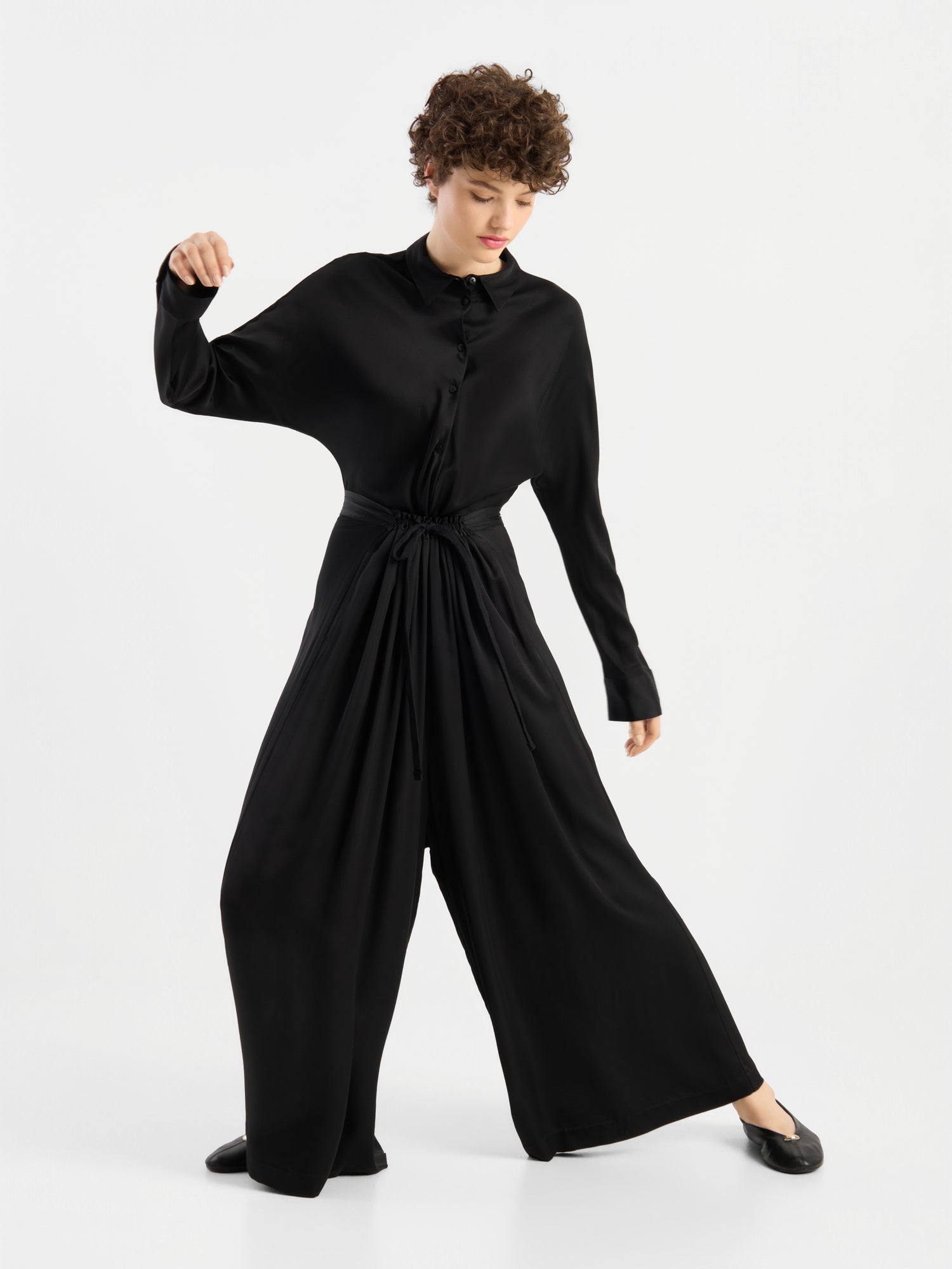 Eine Person mit kurzen Locken, die die Bluse „Kaia Schwarz“ – eine locker sitzende schwarze Bluse – trägt, kombiniert mit schwarzen Schuhen, steht vor einem weißen Hintergrund in einer dynamischen Pose, wobei ein Arm angewinkelt und der andere ausgestreckt ist. Die Bluse wird in Portugal hergestellt und zeugt von hochwertiger Handwerkskunst und zeitgemäßem Stil.