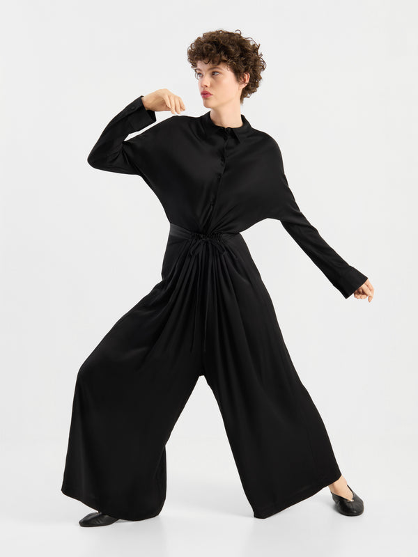 Ein Model trägt „The Art of Black“, einen locker sitzenden schwarzen Jumpsuit mit weitem Bein, und steht in einer dynamischen Pose vor einem schlichten weißen Hintergrund.