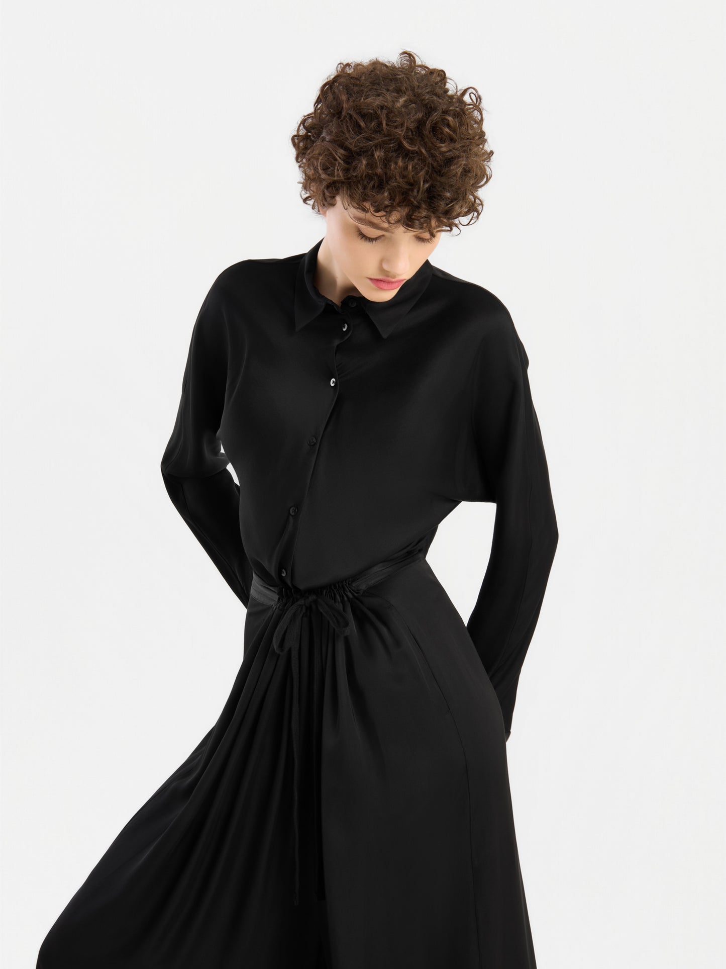 Eine Person mit kurzen Locken, die das Kleid „Bluse Kaia Schwarz“ – ein langärmeliges Kleid mit Knopfleiste – trägt, blickt nach unten, während sie vor einem schlichten weißen Hintergrund steht. Dieses elegante Kleidungsstück erinnert an den lässigen Stil einer fließenden Bluse und wird in Portugal hergestellt.