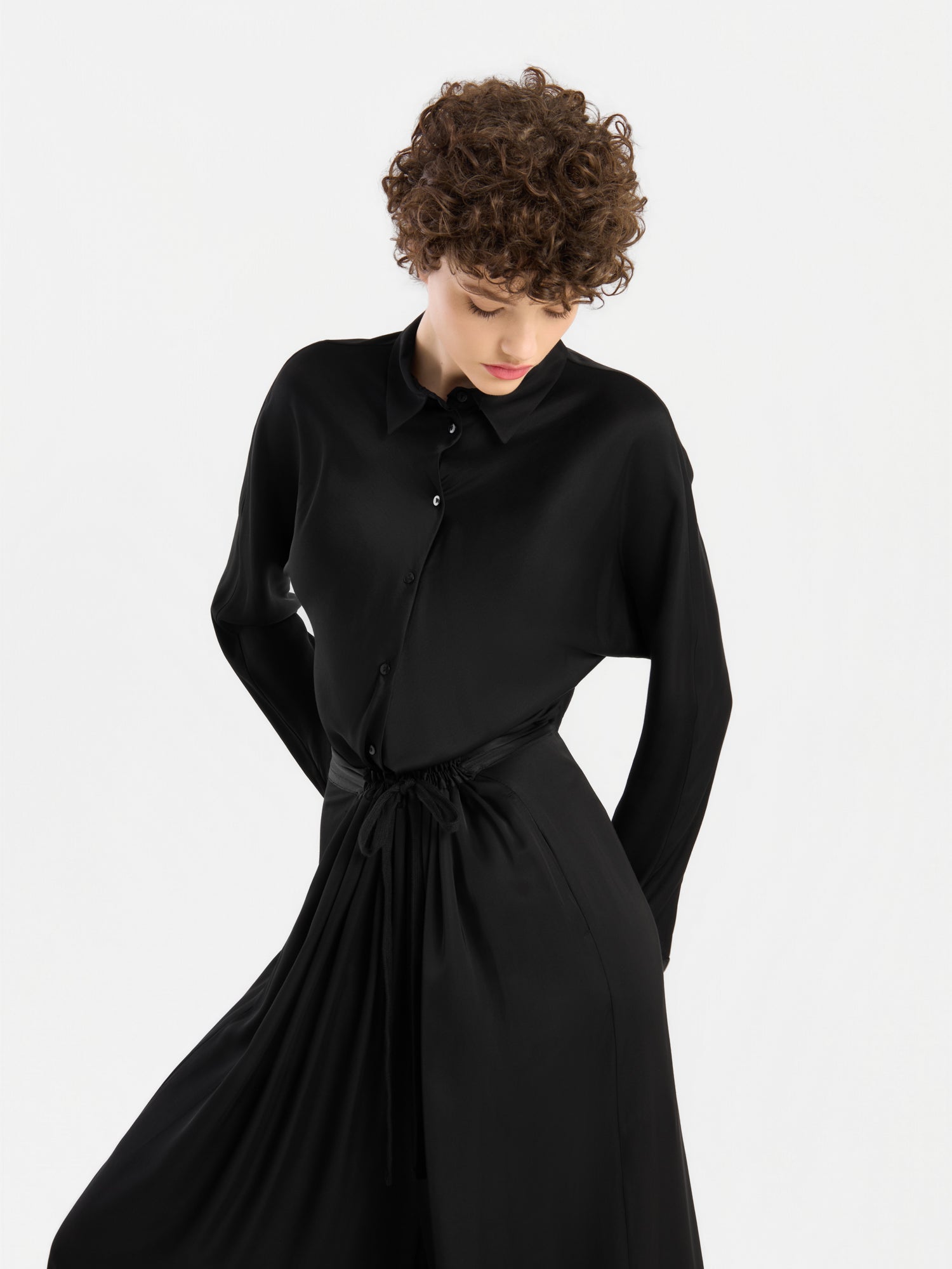 Eine Person mit kurzen Locken, die das Kleid „Bluse Kaia Schwarz“ – ein langärmeliges Kleid mit Knopfleiste – trägt, blickt nach unten, während sie vor einem schlichten weißen Hintergrund steht. Dieses elegante Kleidungsstück erinnert an den lässigen Stil einer fließenden Bluse und wird in Portugal hergestellt.