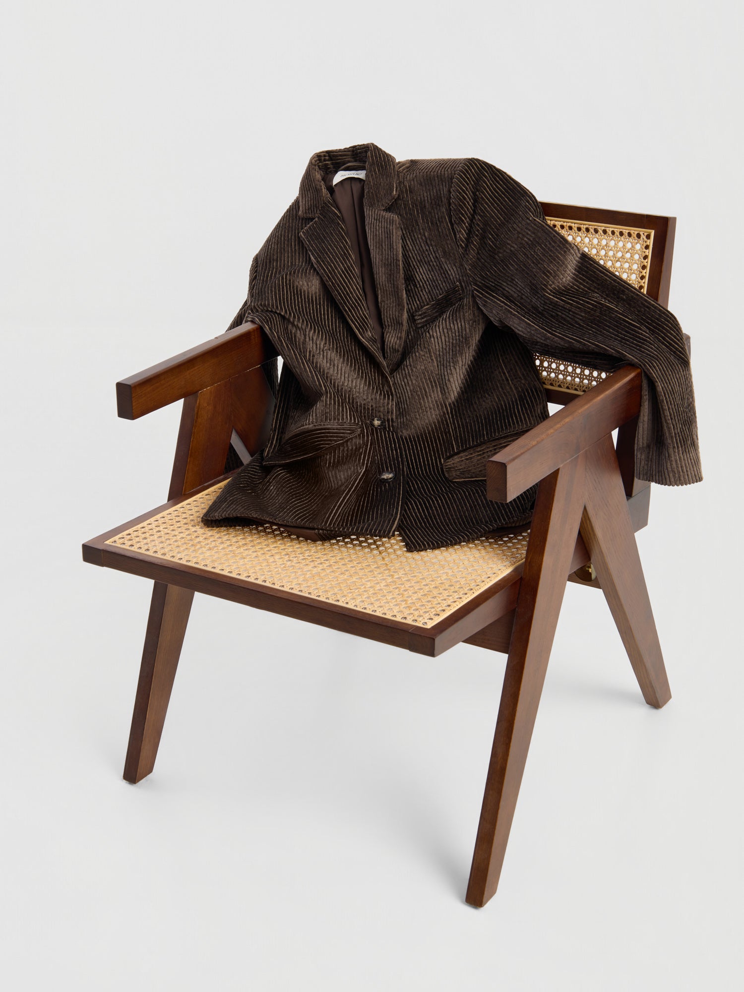 Der Blazer „Alice“ aus braunem Cord liegt über der Rückenlehne und der Sitzfläche eines Holzsessels mit geflochtener Sitzfläche und Rückenlehne aus Rattan und ist vor einem schlichten weißen Hintergrund zu sehen.