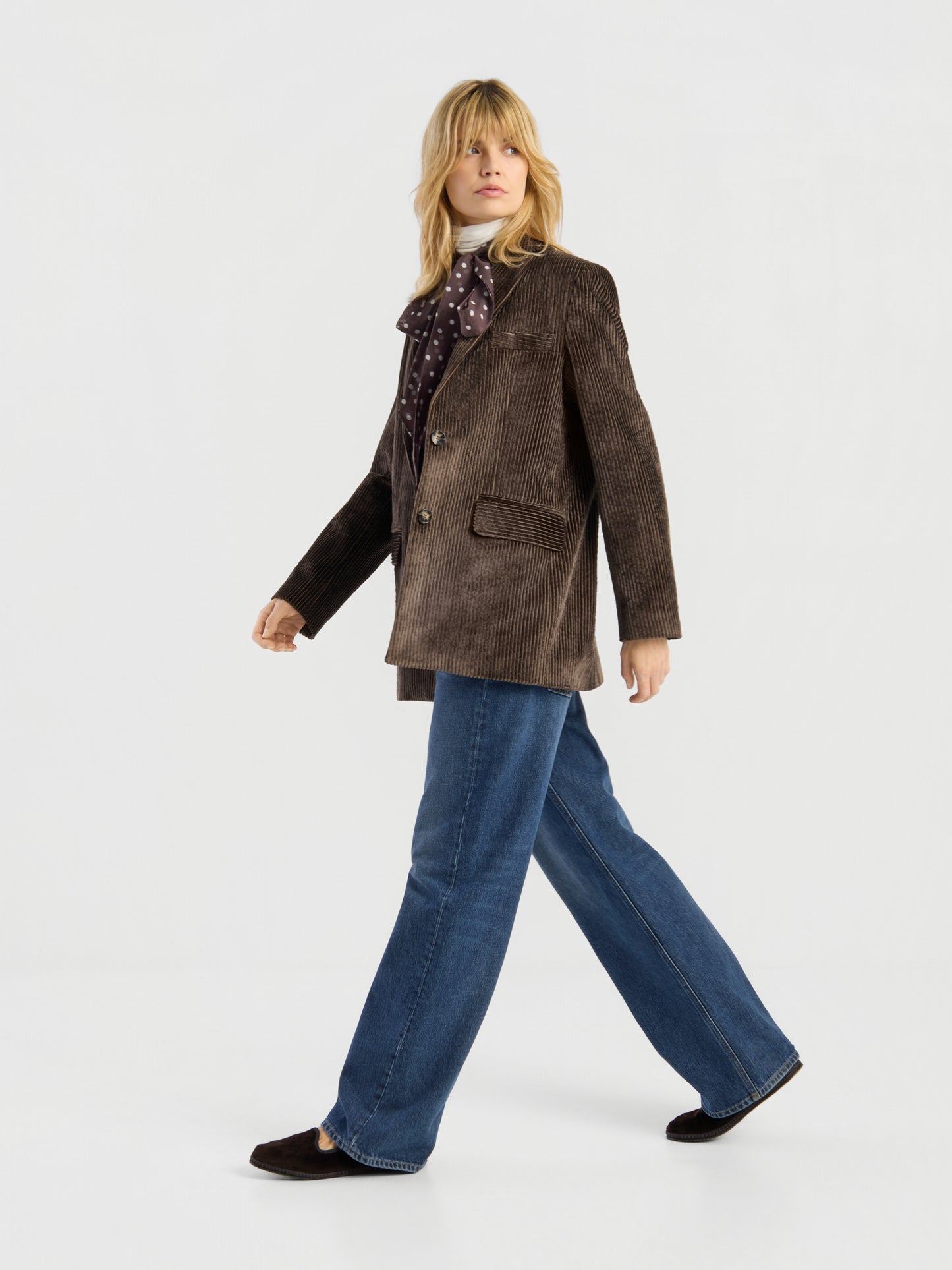 Eine Frau trägt den Blazer „Alice“ aus braunem Cord und kombiniert ihn mit einer blauen Jeans mit weitem Bein sowie dunklen Schuhen, während sie selbstbewusst vor einem schlichten weißen Hintergrund entlanggeht.