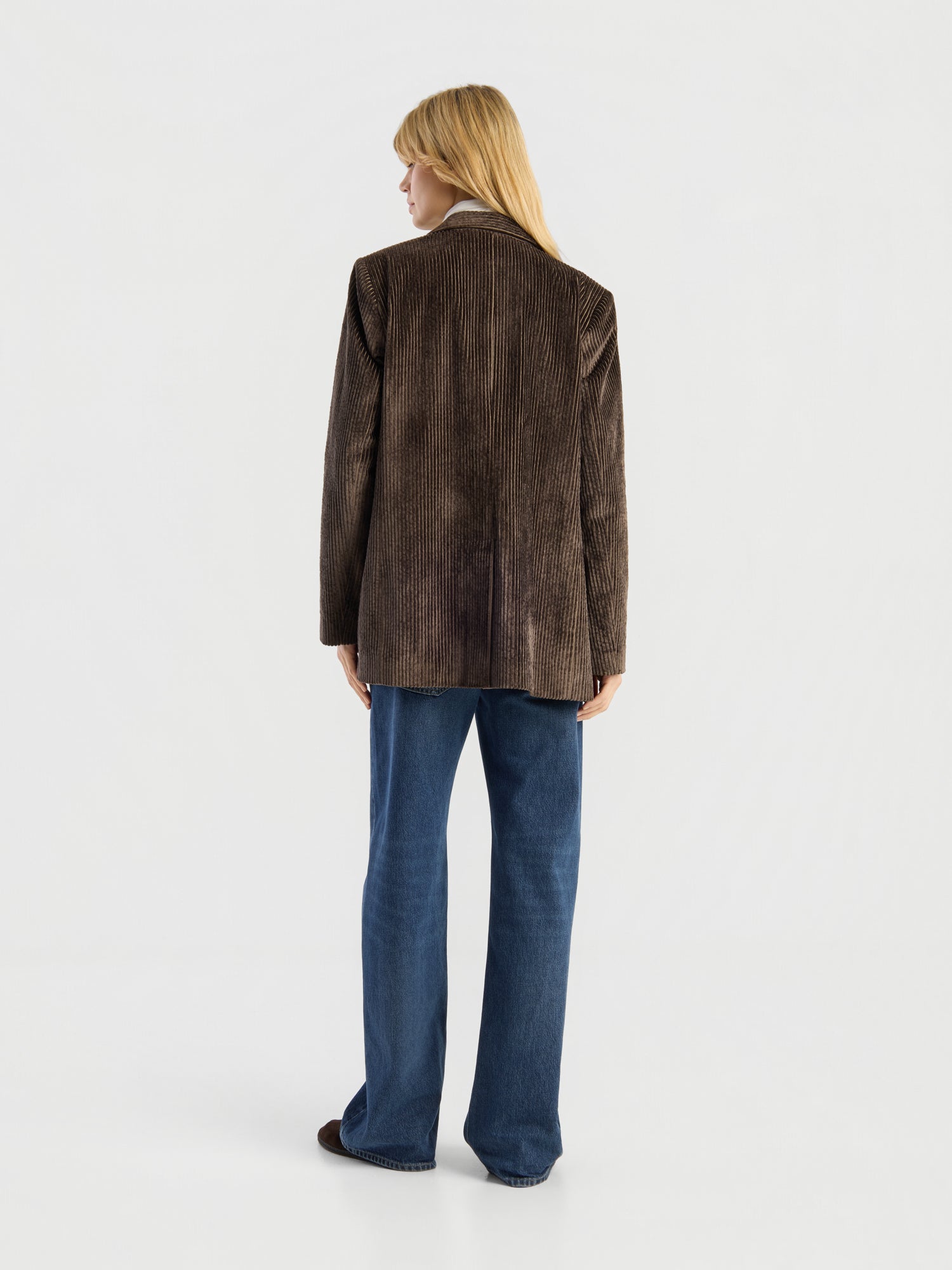 Eine Person steht mit dem Rücken zur Kamera und trägt den Blazer „Alice“ aus braunem Cord sowie eine blaue Jeans. Der Hintergrund ist einfarbig weiß.