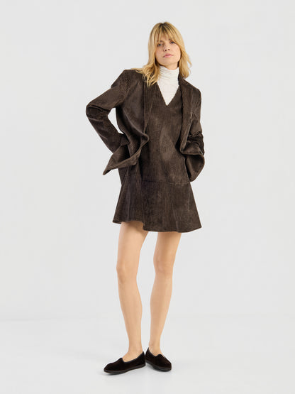 Vor einem schlichten, hellen Hintergrund steht eine Frau, die das „Kleid Alice“ trägt – ein braunes Baumwollkleid aus Cord, das als Set aus Blazer und passendem Rock gestaltet ist. Dazu kombiniert sie einen weißen Rollkragenpullover und schwarze flache Schuhe. Mit den Händen in den Jackentaschen blickt sie nach vorne und verkörpert damit zeitgenössische portugiesische Mode, die vom klassischen „Damen Kleid Portugal“ inspiriert ist.
