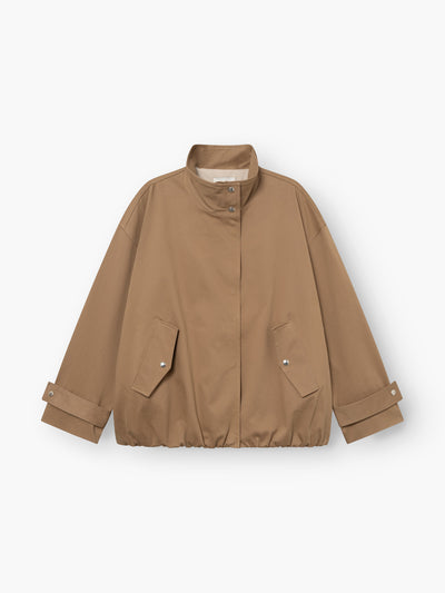 Eine hellbraune, locker sitzende „Jacke Hamptons“ – eine Damen-Übergangsjacke mit Stehkragen, Druckknopfleiste vorne, zwei Pattentaschen und verstellbaren Ärmelbündchen. Der Saum ist leicht gerafft. Diese im Utility-Stil gehaltene Jacke ist flach auf einem weißen Hintergrund ausgelegt.