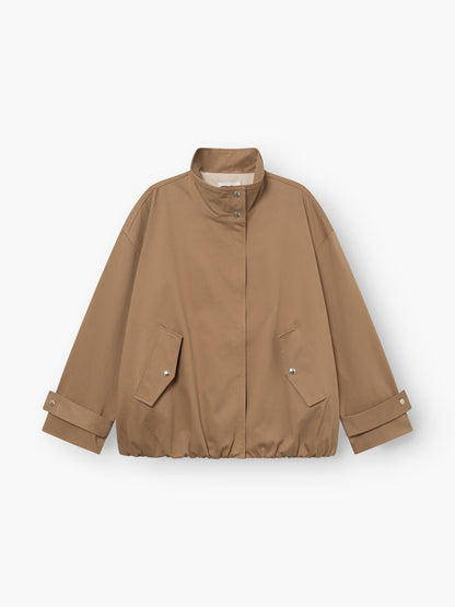 Eine hellbraune, locker sitzende „Jacke Hamptons“ – eine Damen-Übergangsjacke mit Stehkragen, Druckknopfleiste vorne, zwei Pattentaschen und verstellbaren Ärmelbündchen. Der Saum ist leicht gerafft. Diese im Utility-Stil gehaltene Jacke ist flach auf einem weißen Hintergrund ausgelegt.