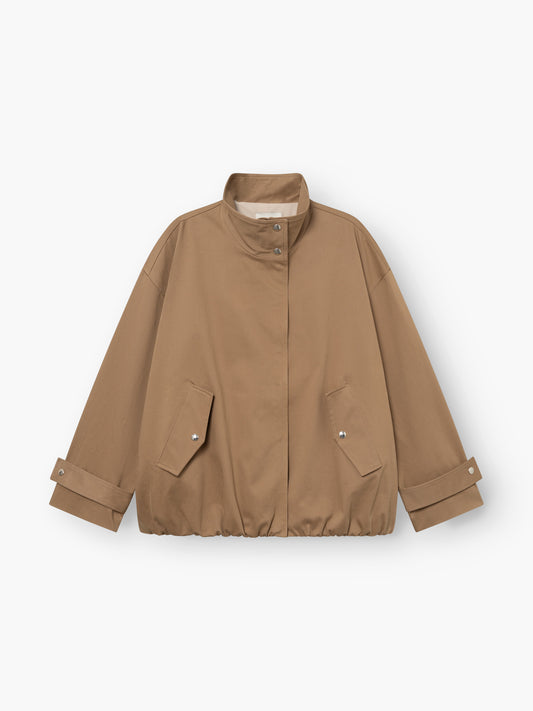 Eine hellbraune, locker sitzende „Jacke Hamptons“ – eine Damen-Übergangsjacke mit Stehkragen, Druckknopfleiste vorne, zwei Pattentaschen und verstellbaren Ärmelbündchen. Der Saum ist leicht gerafft. Diese im Utility-Stil gehaltene Jacke ist flach auf einem weißen Hintergrund ausgelegt.