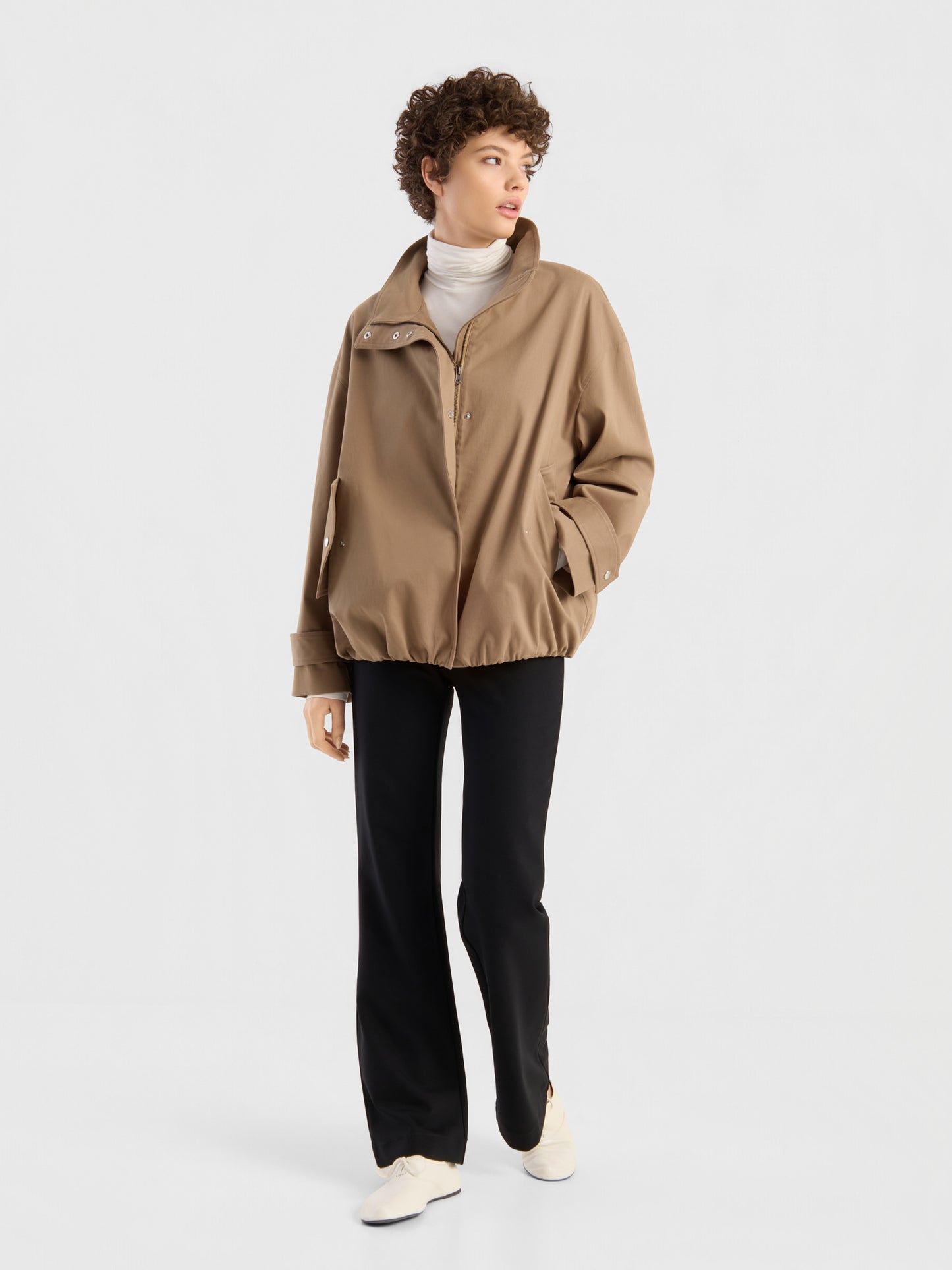 Eine Person, die eine hellbraune, übergroße Jacke der Marke „Hamptons“, einen weißen Rollkragenpullover, eine schwarze Hose und weiße Schuhe trägt, steht vor einem schlichten weißen Hintergrund und blickt zur Seite.