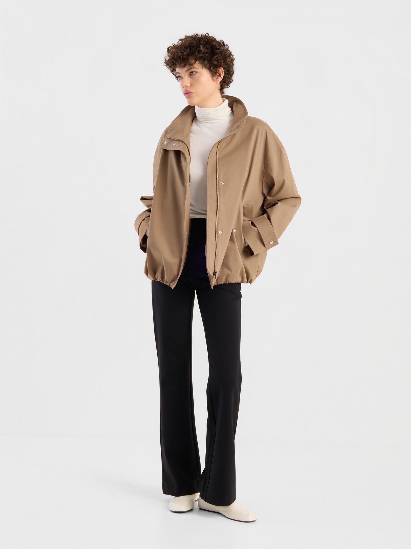 Eine Person steht vor einem schlichten Hintergrund und trägt eine hellbraune, übergroße Jacke im Hamptons-Stil, die als Damen-Übergangsjacke gestylt ist, dazu einen weißen Rollkragenpullover, eine schwarze Schlaghose und weiße Schuhe; die Hände hat sie lässig in die Jackentaschen gesteckt.