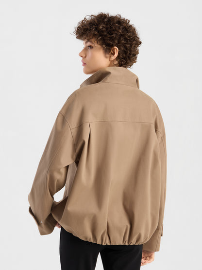 Eine Person mit kurzen Locken trägt die übergroße Jacke „Hamptons“ in Beige mit hohem Kragen, während sie mit dem Rücken zur Kamera steht und vor einem schlichten weißen Hintergrund über die Schulter blickt.
