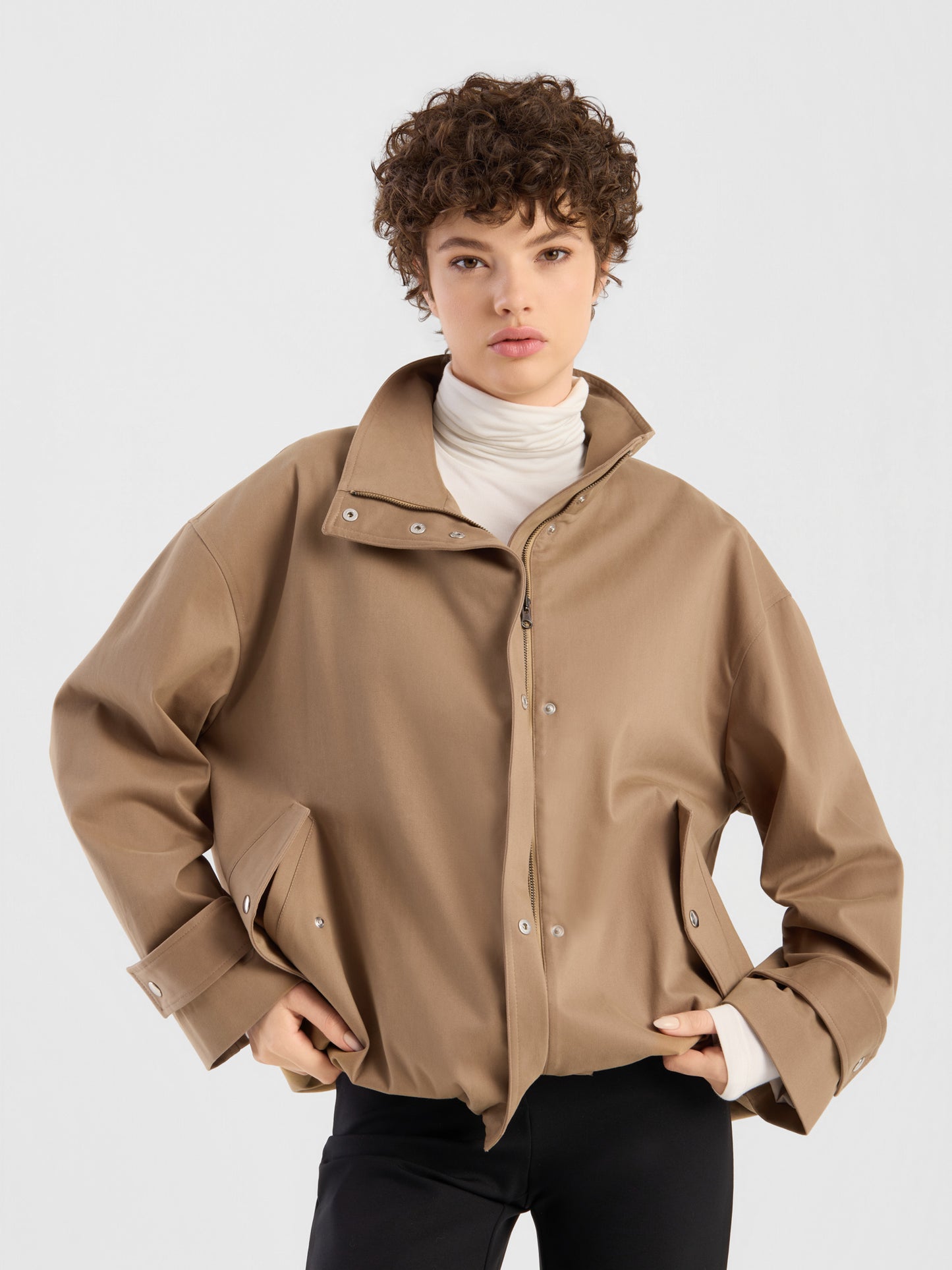 Eine Person mit kurzen Locken trägt die Oversize-Jacke „Hamptons“ mit breiten Ärmelbündchen über einem weißen Rollkragenpullover und einer schwarzen Hose und steht mit den Händen in den Hüften vor einem schlichten, hellen Hintergrund.
