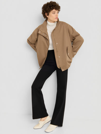 Eine Person mit kurzen Locken trägt eine übergroße Jacke von „Hamptons“ über einem weißen Rollkragenpullover, dazu eine schwarze Schlaghose und weiße Schuhe, und steht vor einer schlichten, hellen Wand.