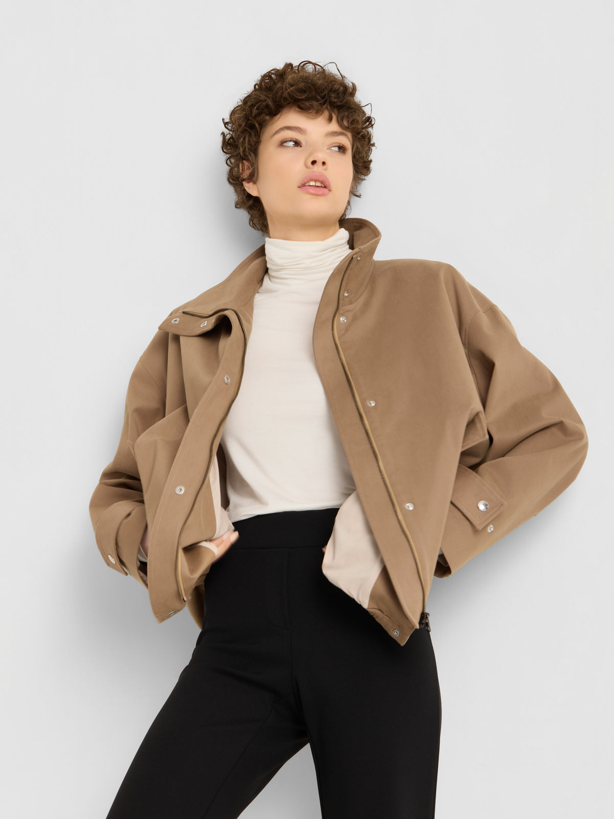 Eine Person, die eine hellbraune Jacke über einem weißen Rollkragenpullover und die „Travel Pants“ in Schwarz trägt, steht mit den Händen in den Taschen und blickt vor einem schlichten, hellen Hintergrund zur Seite. Diese Damenhose mit ausgestelltem Bein, hergestellt in Portugal, rundet den mühelos stilvollen Look ab.