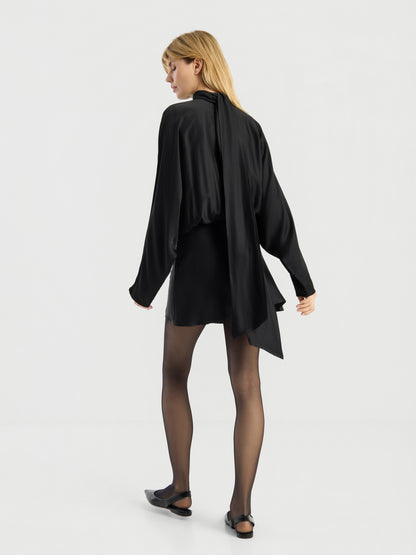 Eine Frau steht vor einem schlichten weißen Hintergrund, den Rücken zur Kamera gewandt und den Blick leicht zur Seite gerichtet. Sie trägt das „Dress Antonia Bow“ – ein schwarzes, langärmeliges Kleid mit einem Schal-Detail –, dazu schwarze, transparente Strumpfhosen und schwarze Ballerinas.