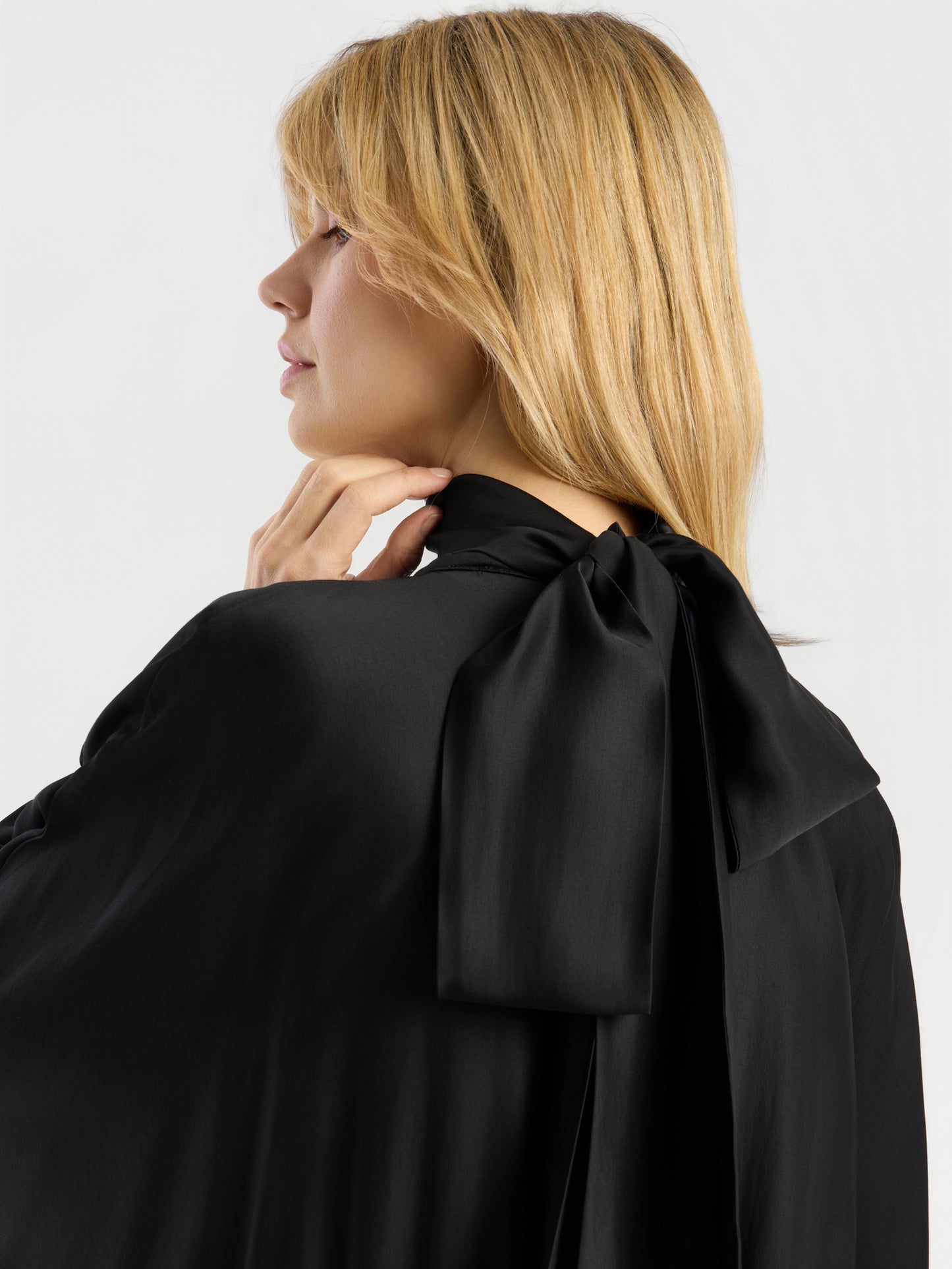Eine Frau mit blonden Haaren trägt das Kleid „Antonia Bow“, das sich durch eine große, seidige Schleife auszeichnet, die im Nacken gebunden ist; sie berührt sanft ihr Kinn und blickt zur Seite.
