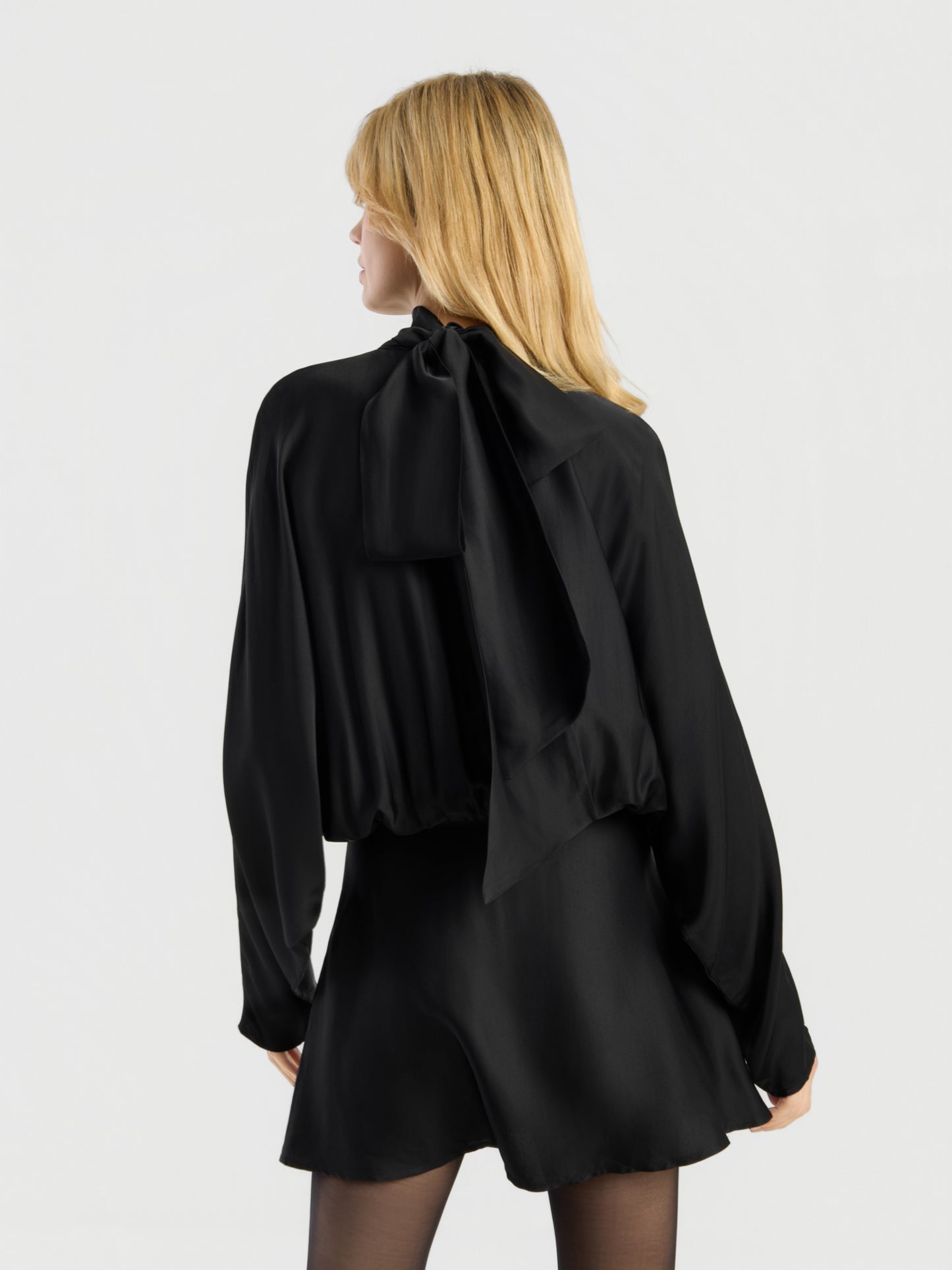 Eine Person mit blonden Haaren steht vor einem schlichten, hellen Hintergrund, mit dem Rücken zur Kamera, und trägt das „Dress Antonia Bow“ – eine schwarze, langärmelige Bluse mit einer großen Schleife am Halsausschnitt und einen dazu passenden schwarzen Rock.