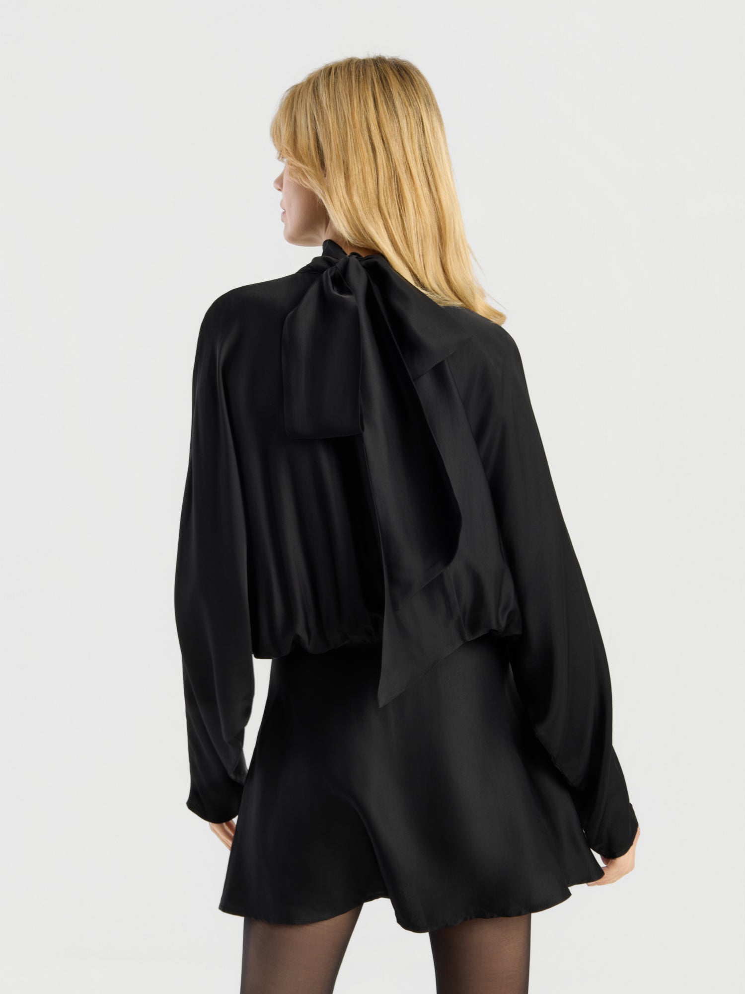 Eine Person mit blonden Haaren steht vor einem schlichten, hellen Hintergrund, mit dem Rücken zur Kamera, und trägt das „Dress Antonia Bow“ – eine schwarze, langärmelige Bluse mit einer großen Schleife am Halsausschnitt und einen dazu passenden schwarzen Rock.