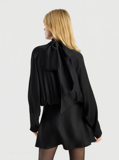 Eine Person mit blonden Haaren steht vor einem schlichten, hellen Hintergrund, mit dem Rücken zur Kamera, und trägt das „Dress Antonia Bow“ – eine schwarze, langärmelige Bluse mit einer großen Schleife am Halsausschnitt und einen dazu passenden schwarzen Rock.