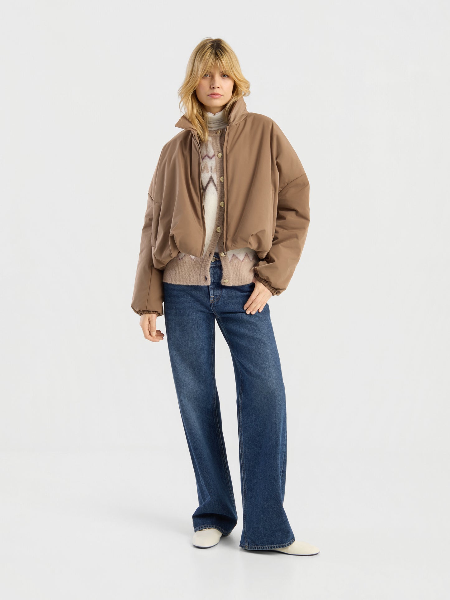 Eine Person, die die Ballon-Jacke in Beige trägt, kombiniert mit einem gemusterten Pullover, blauen Jeans mit weitem Bein und weißen Schuhen, steht vor einem schlichten weißen Hintergrund.