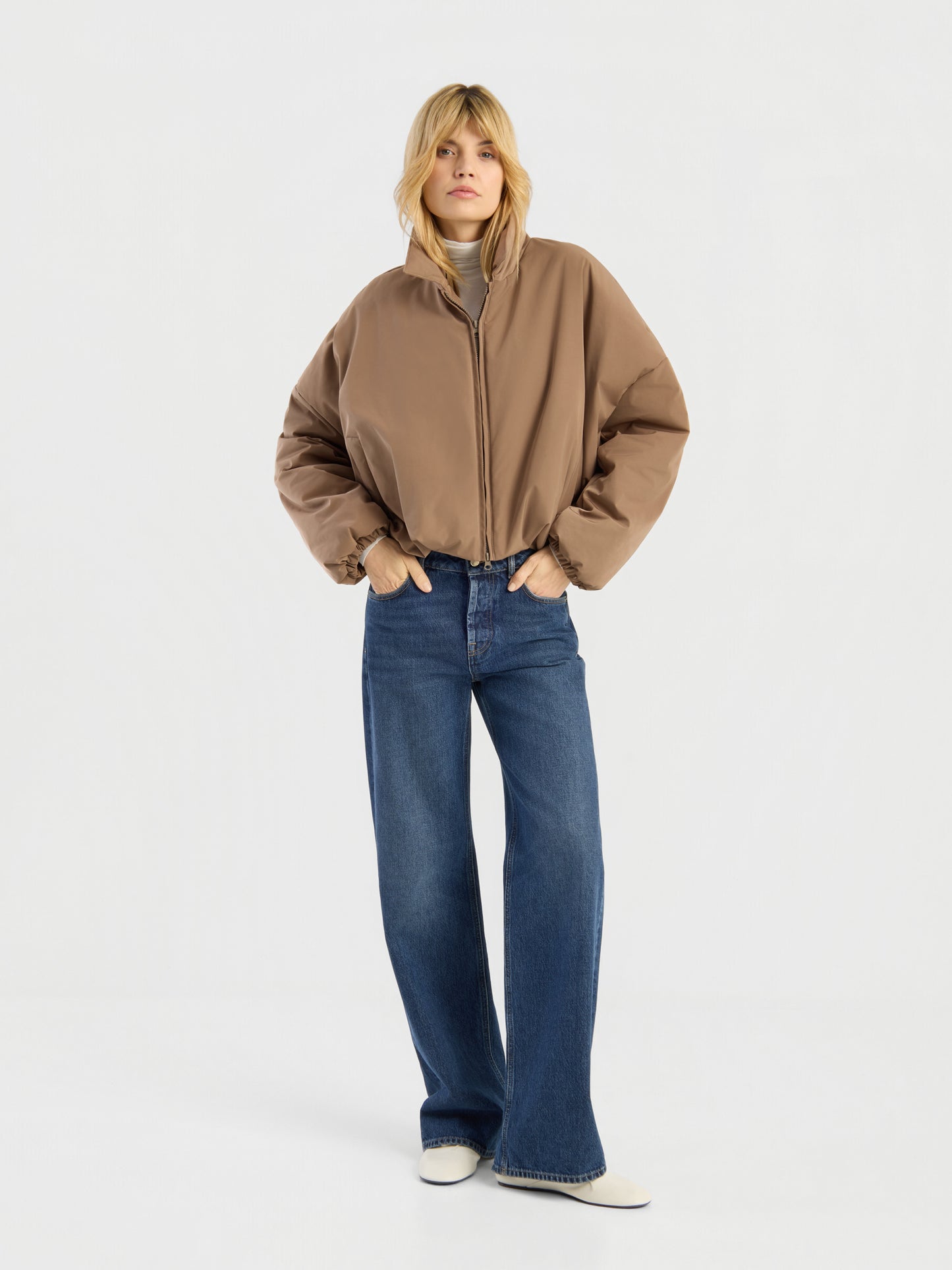 Eine Person, die eine Ballon-Jacke, blaue Jeans mit weitem Bein und weiße Schuhe trägt, steht mit den Händen in den Taschen vor einem schlichten weißen Hintergrund.