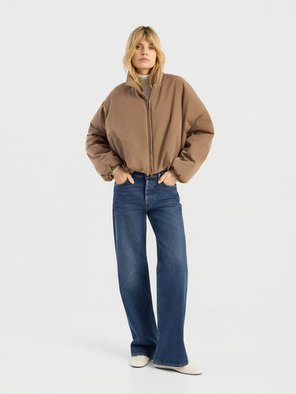 Eine Person, die eine Ballon-Jacke, blaue Jeans mit weitem Bein und weiße Schuhe trägt, steht mit den Händen in den Taschen vor einem schlichten weißen Hintergrund.