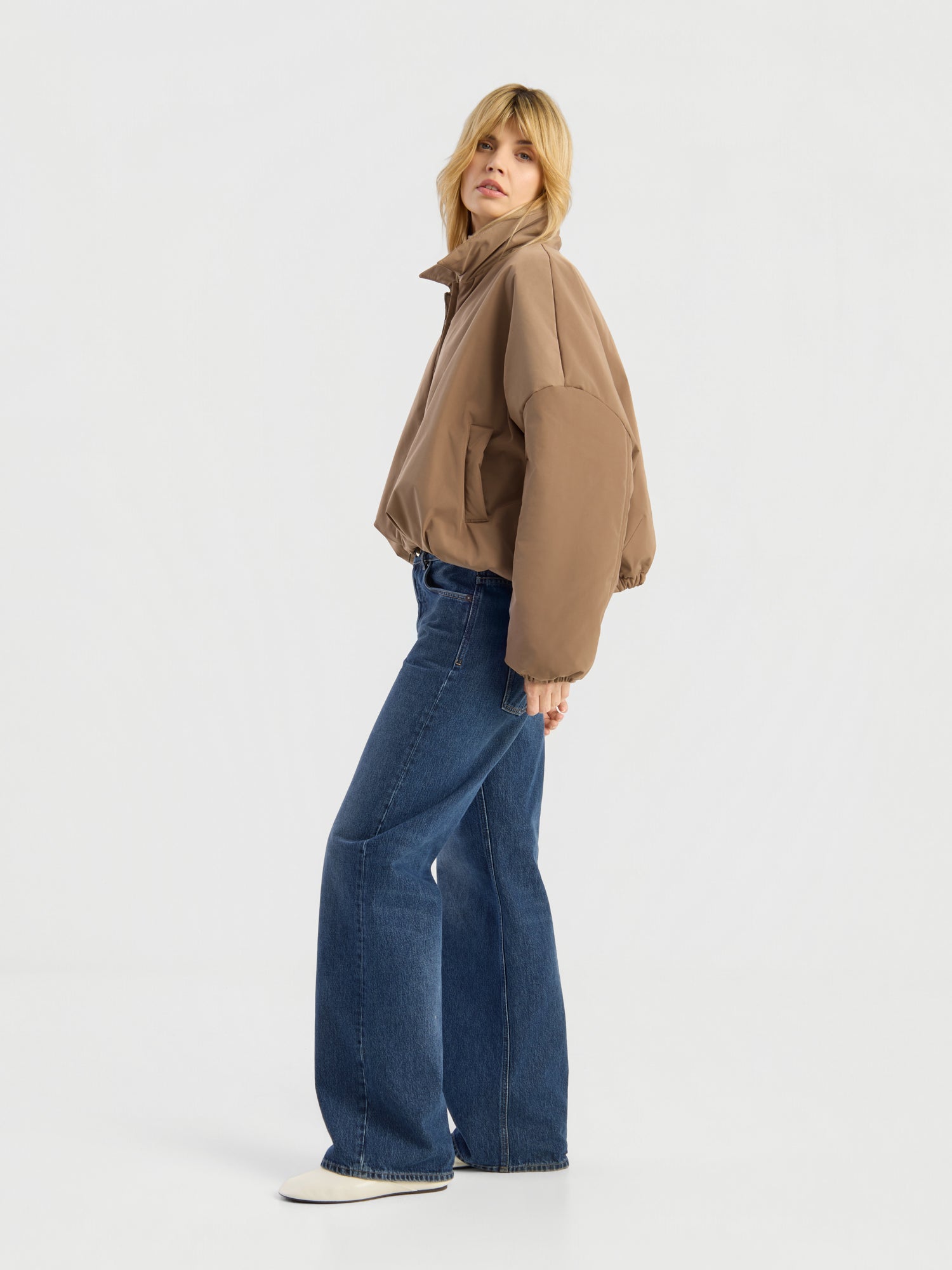 Eine Person mit blonden Haaren trägt die Ballon-Jacke in Beige, eine blaue Jeans mit weitem Bein und weiße Schuhe und steht seitlich vor einem schlichten weißen Hintergrund.
