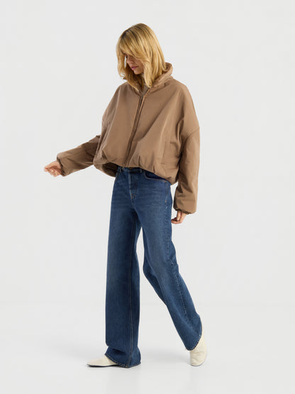 Eine Person mit blonden Haaren trägt die Ballon-Jacke in Braun, dazu eine blaue Jeans mit weitem Bein und weiße Schuhe, und steht vor einem schlichten weißen Hintergrund.