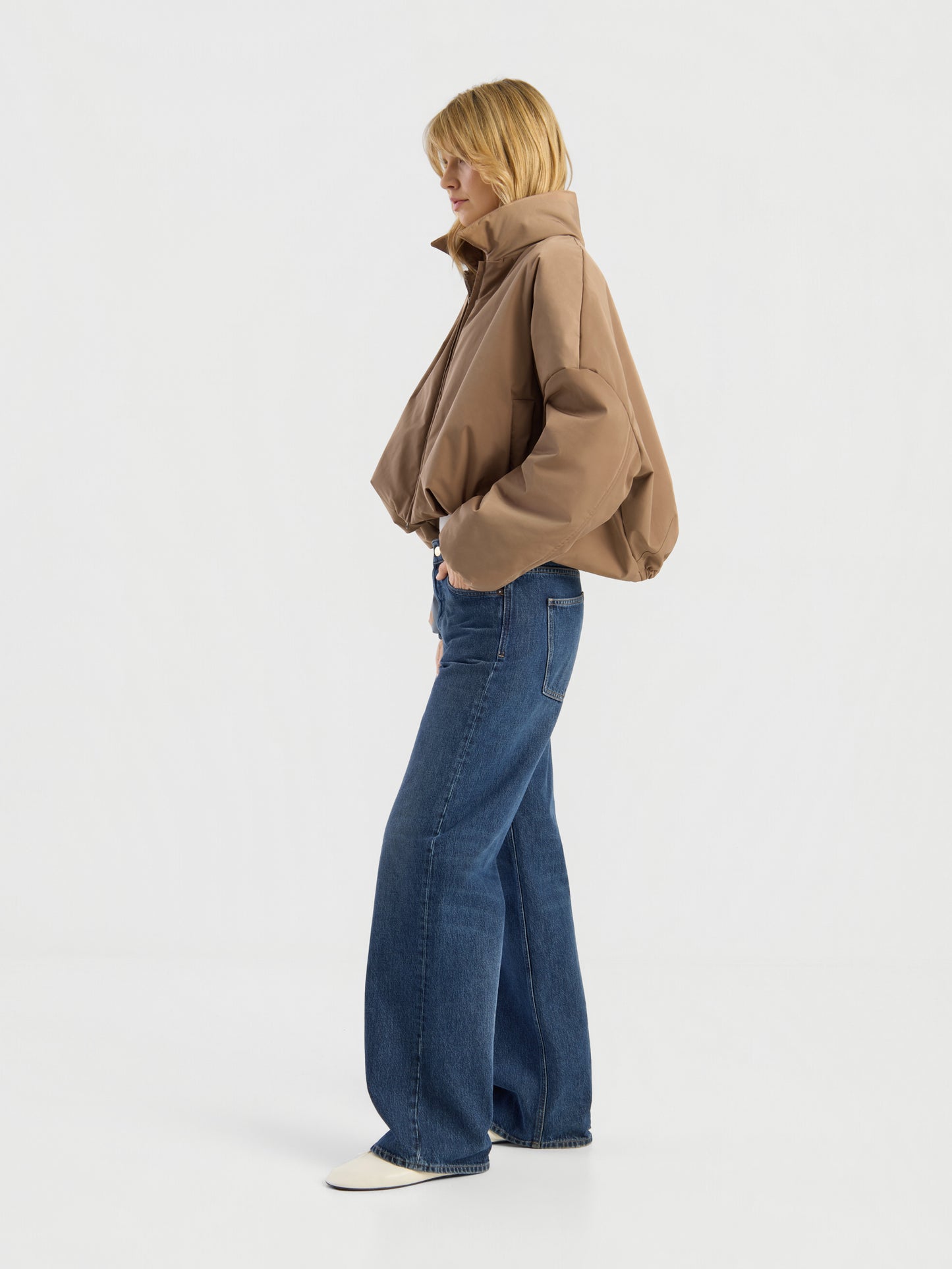 Eine Person, die eine Ballon-Jacke, blaue Jeans mit weitem Bein und weiße Schuhe trägt, steht seitlich vor einem schlichten weißen Hintergrund.