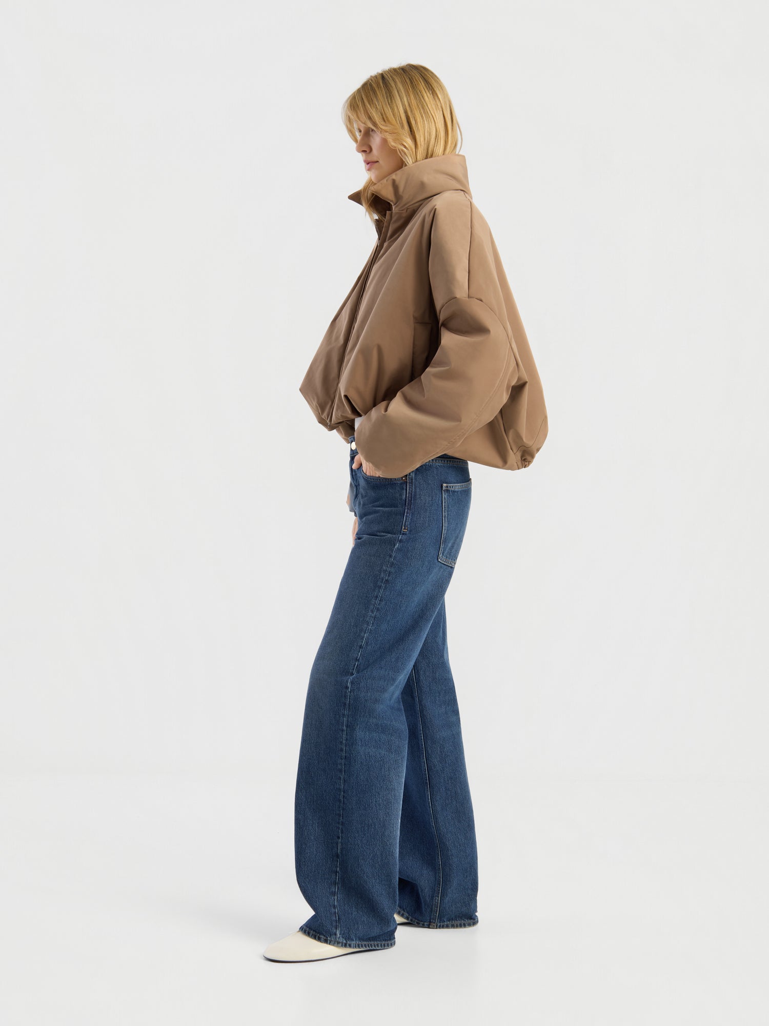 Eine Person, die eine Ballon-Jacke, blaue Jeans mit weitem Bein und weiße Schuhe trägt, steht seitlich vor einem schlichten weißen Hintergrund.