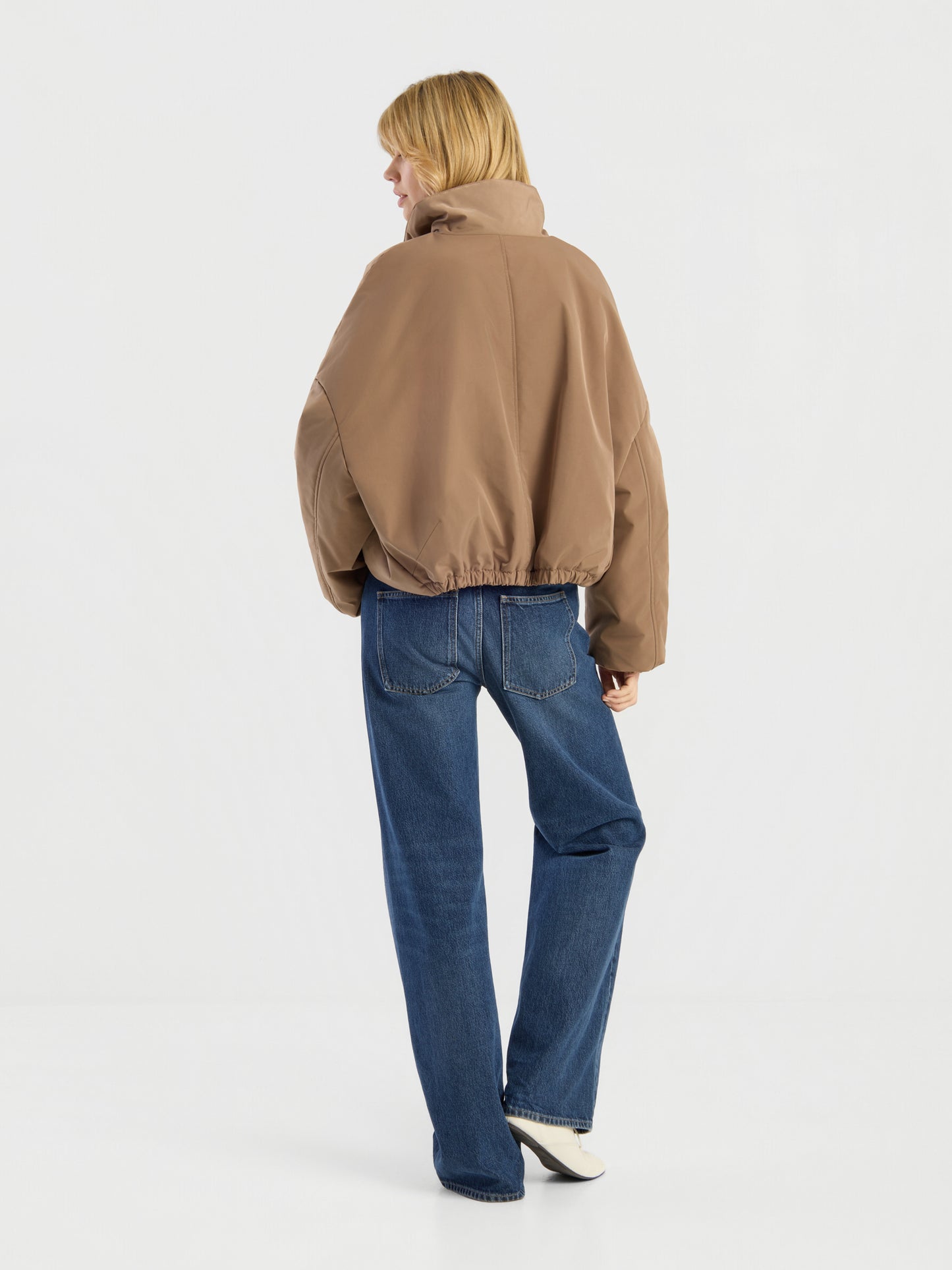 Eine Person mit blonden Haaren, die eine braune Ballonjacke, blaue Jeans und weiße Schuhe trägt, steht mit dem Rücken zur Kamera vor einem schlichten weißen Hintergrund.