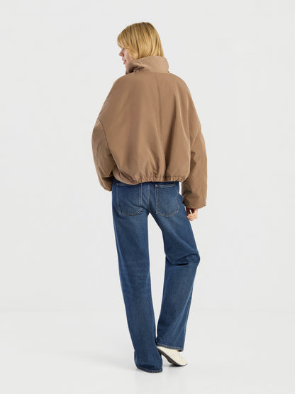 Eine Person mit blonden Haaren, die eine braune Ballonjacke, blaue Jeans und weiße Schuhe trägt, steht mit dem Rücken zur Kamera vor einem schlichten weißen Hintergrund.