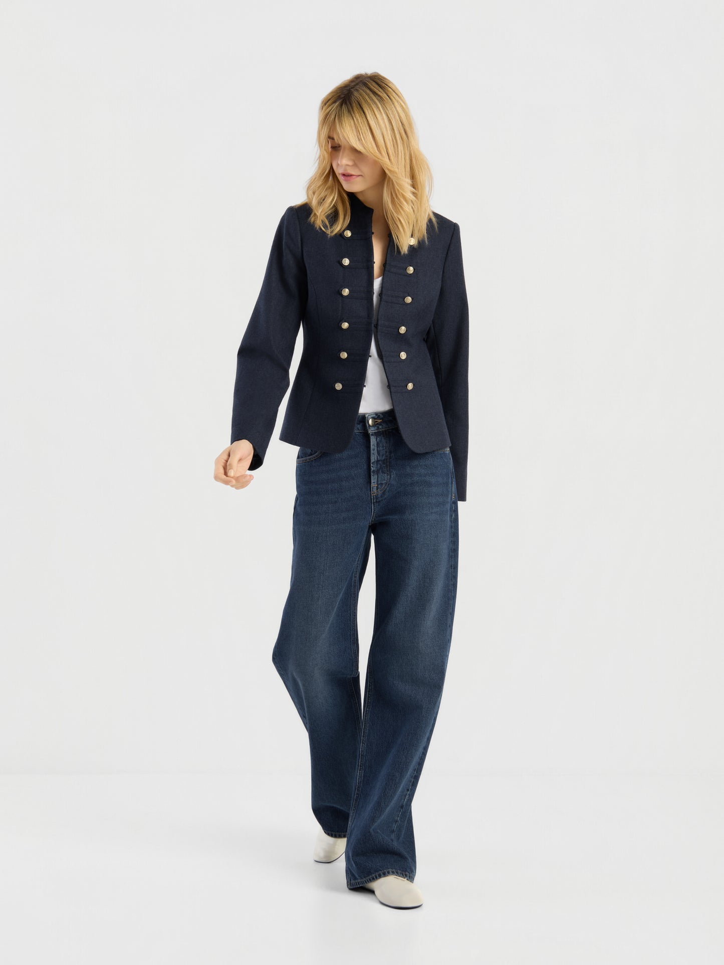 Eine Frau mit schulterlangen blonden Haaren, die die marineblaue Jacke „Amelia“ im Military-Stil mit goldenen Knöpfen, ein weißes Hemd, eine blaue Jeans mit weitem Bein und weiße Schuhe trägt und vor einem schlichten hellen Hintergrund steht.