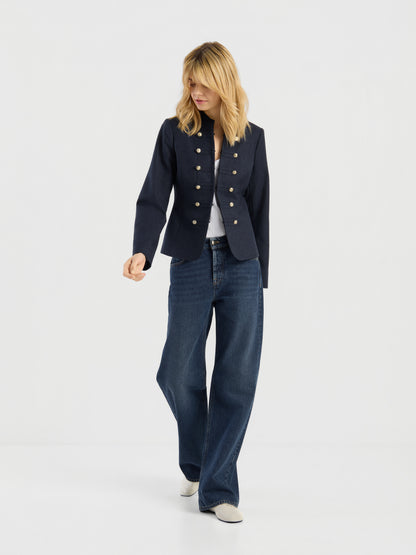Eine Frau mit schulterlangen blonden Haaren, die die marineblaue Jacke „Amelia“ im Military-Stil mit goldenen Knöpfen, ein weißes Hemd, eine blaue Jeans mit weitem Bein und weiße Schuhe trägt und vor einem schlichten hellen Hintergrund steht.