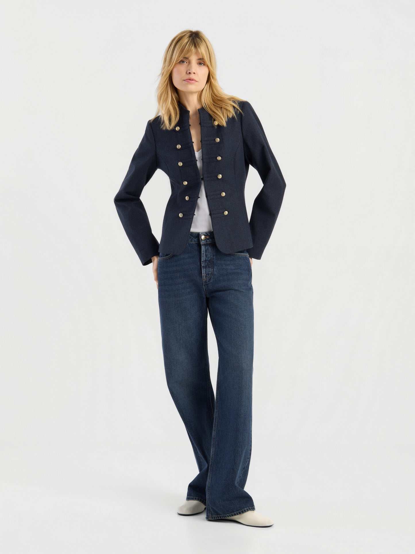 Eine Person, die eine marineblaue Jacke „Amelia“ im Military-Stil mit goldenen Knöpfen, ein weißes Oberteil, eine blaue Jeans mit weitem Bein und weiße Schuhe trägt, steht vor einem schlichten, hellen Hintergrund.