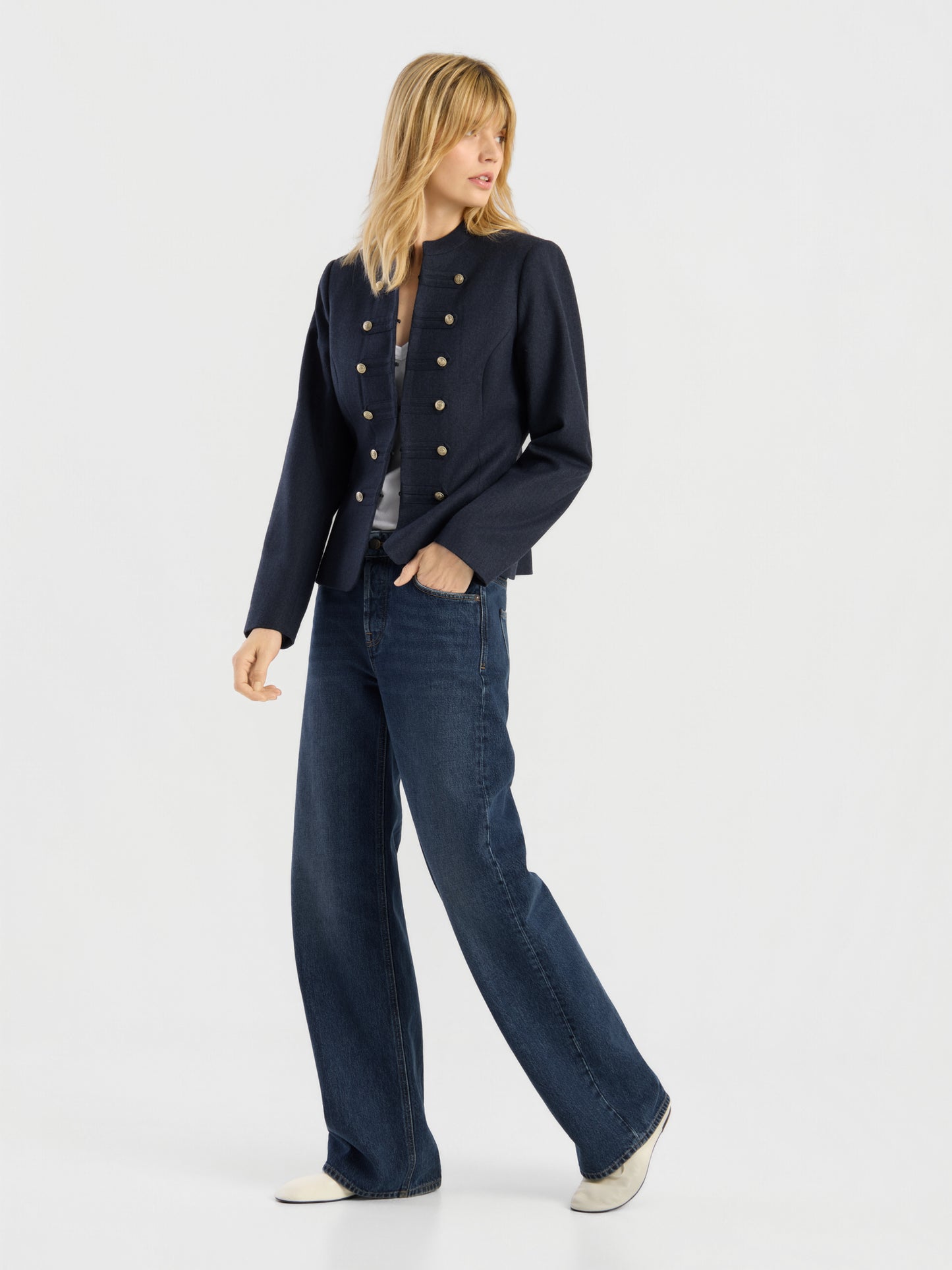 Eine Person, die die Jacke „Amelia“ trägt – eine marineblaue Damenjacke im Military-Stil mit goldenen Knöpfen –, dazu ein graues Hemd, eine blaue Jeans mit weitem Bein und weiße Schuhe, steht vor einem schlichten weißen Hintergrund.