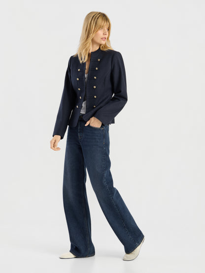 Eine Person, die die Jacke „Amelia“ trägt – eine marineblaue Damenjacke im Military-Stil mit goldenen Knöpfen –, dazu ein graues Hemd, eine blaue Jeans mit weitem Bein und weiße Schuhe, steht vor einem schlichten weißen Hintergrund.
