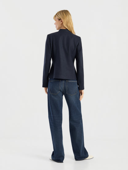 Eine Person mit blonden Haaren, die die dunkle Jacke „Amelia“ im Military-Stil und eine locker sitzende blaue Jeans trägt, steht mit dem Rücken zur Kamera in einem minimalistischen, hellen Studio-Setting.