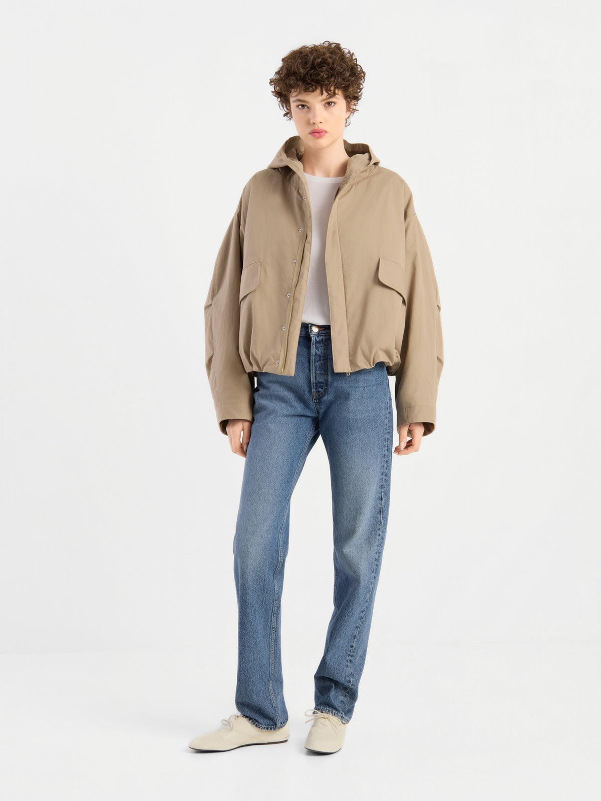 Eine Person mit kurzen Locken trägt die beige Jacke „Savanna“ über einem weißen Oberteil, blauen Jeans und weißen Schuhen und steht vor einem schlichten weißen Hintergrund.