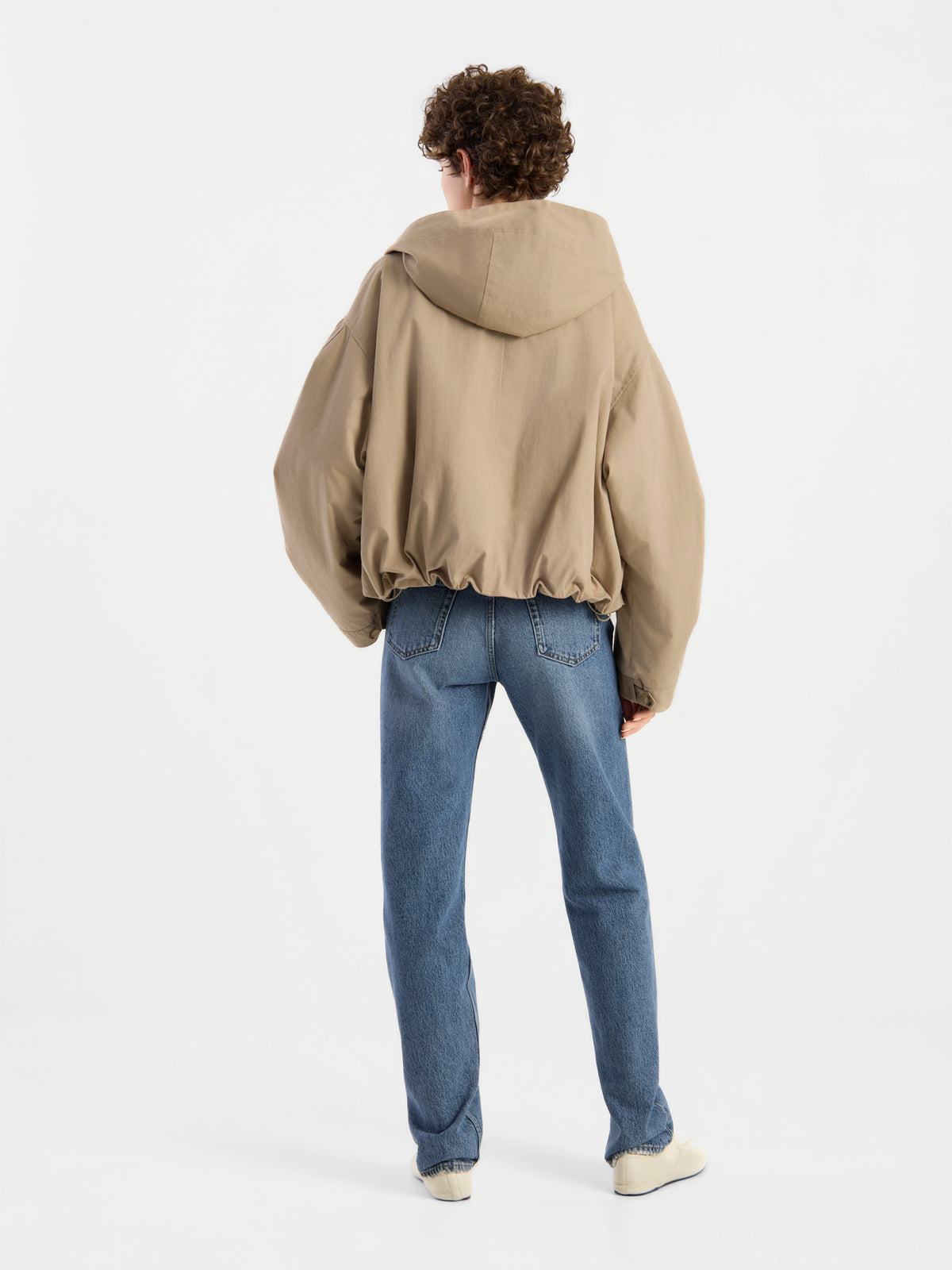 Eine Person mit lockigem Haar trägt die Jacke „Savanna“, eine übergroße beige Damenjacke mit Kapuze, dazu blaue Jeans und weiße Schuhe. Sie steht vor einem schlichten weißen Hintergrund und ist mit dem Rücken zur Kamera gewandt.